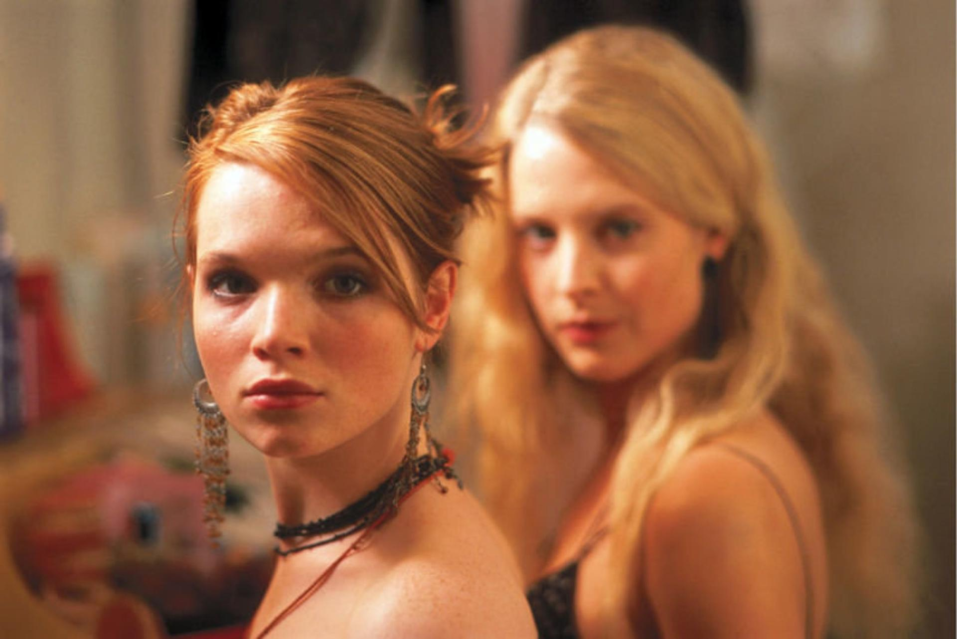 Zwei junge Frauen schauen in die Kamera. Die vordere hat rotes Haar, Ohrringe und eine Halskette. Die andere hat blondes Haar.