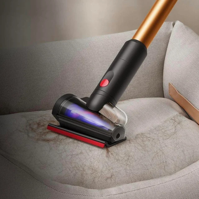 Odkurzacz czyści szarą sofę, usuwając włosy. Odkurzacz jest czarny z czerwonym przyciskiem i czerwonymi akcentami.