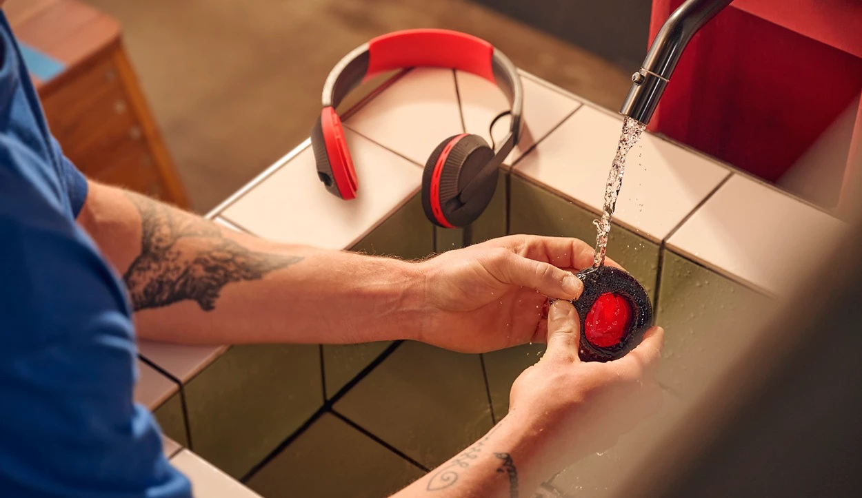 Osoba czyści czerwoną i czarną słuchawkę pod bieżącą wodą. Druga słuchawka i pałąk spoczywają na kafelkowej powierzchni.