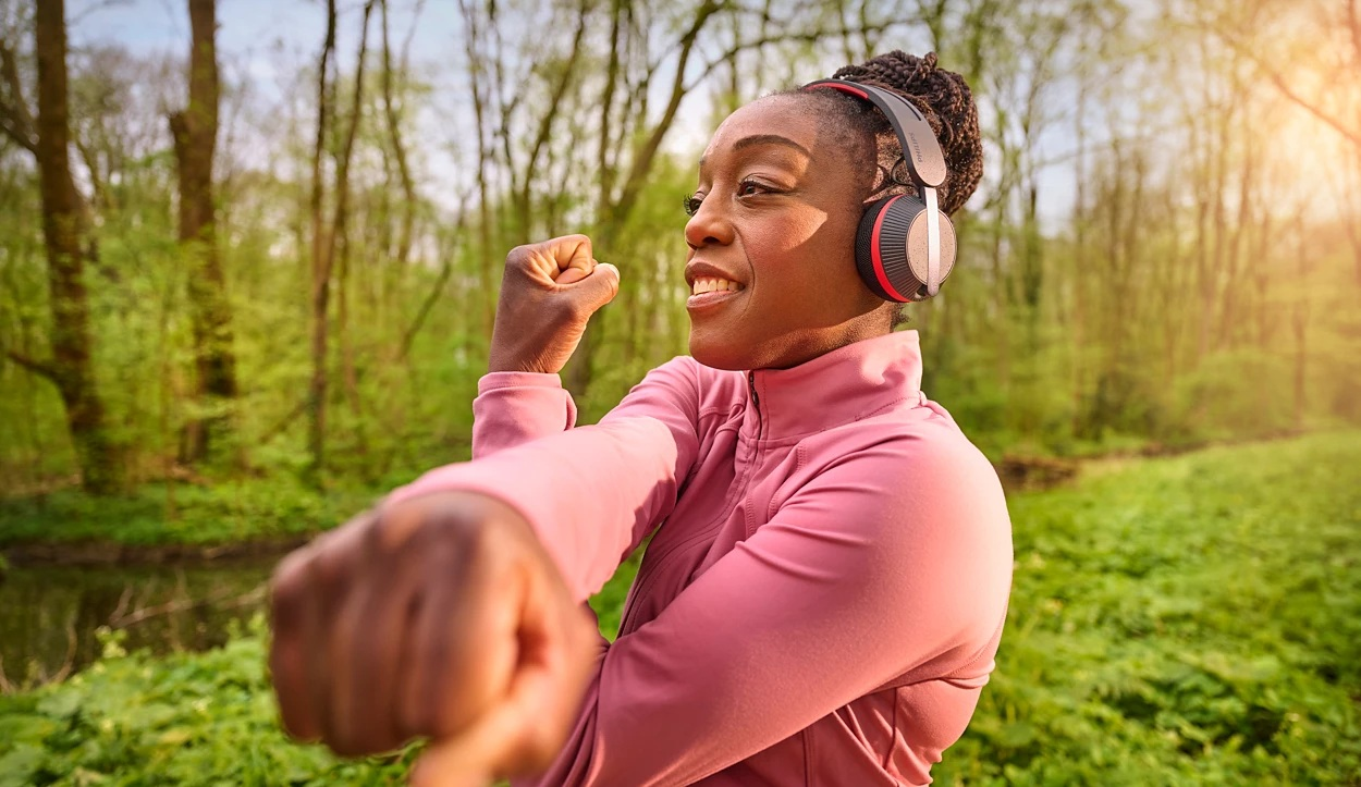 Kobieta w różowej kurtce rozciąga się, w słuchawkach, w lesie z rzeką.