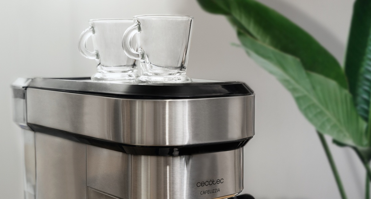 Dwa przezroczyste szklane kubki na ekspresie do kawy ze stali nierdzewnej. Zielone liście po prawej na białym tle.