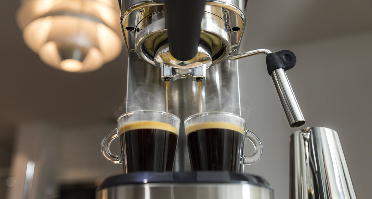 Ekspres do kawy dozuje kawę do dwóch szklanych filiżanek. Para unosi się, lampa jest w tle, a metalowy dzbanek jest z boku.