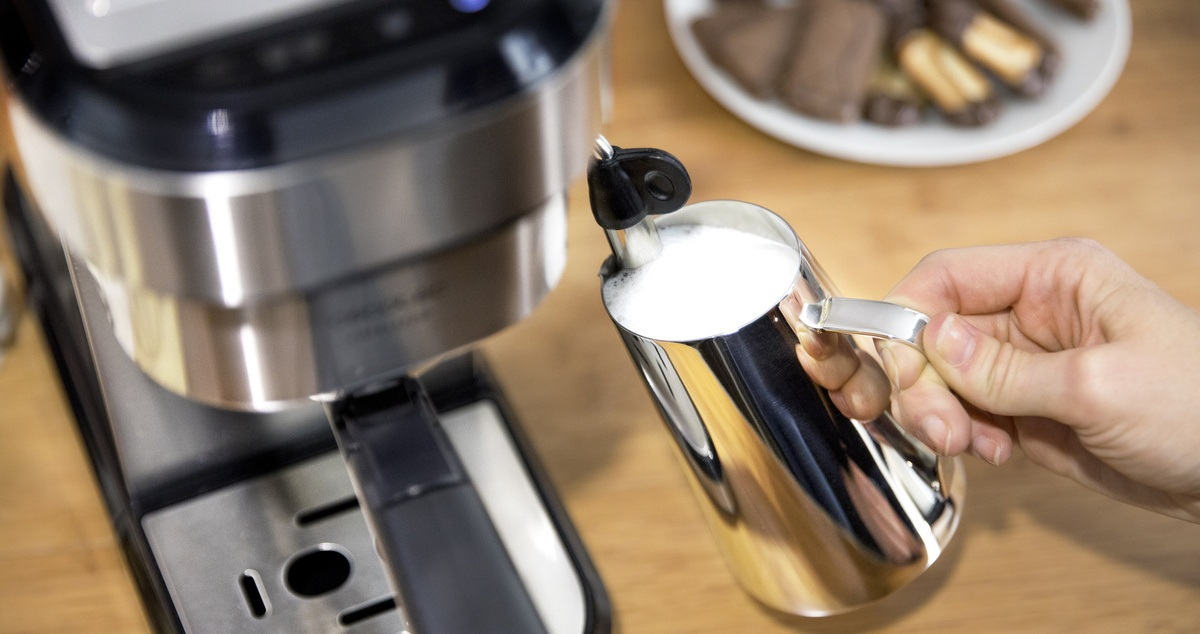 Osoba spienia mleko ekspresem do kawy. Srebrny dzbanek z mlekiem jest z przodu. Ciastka na talerzu w tle.