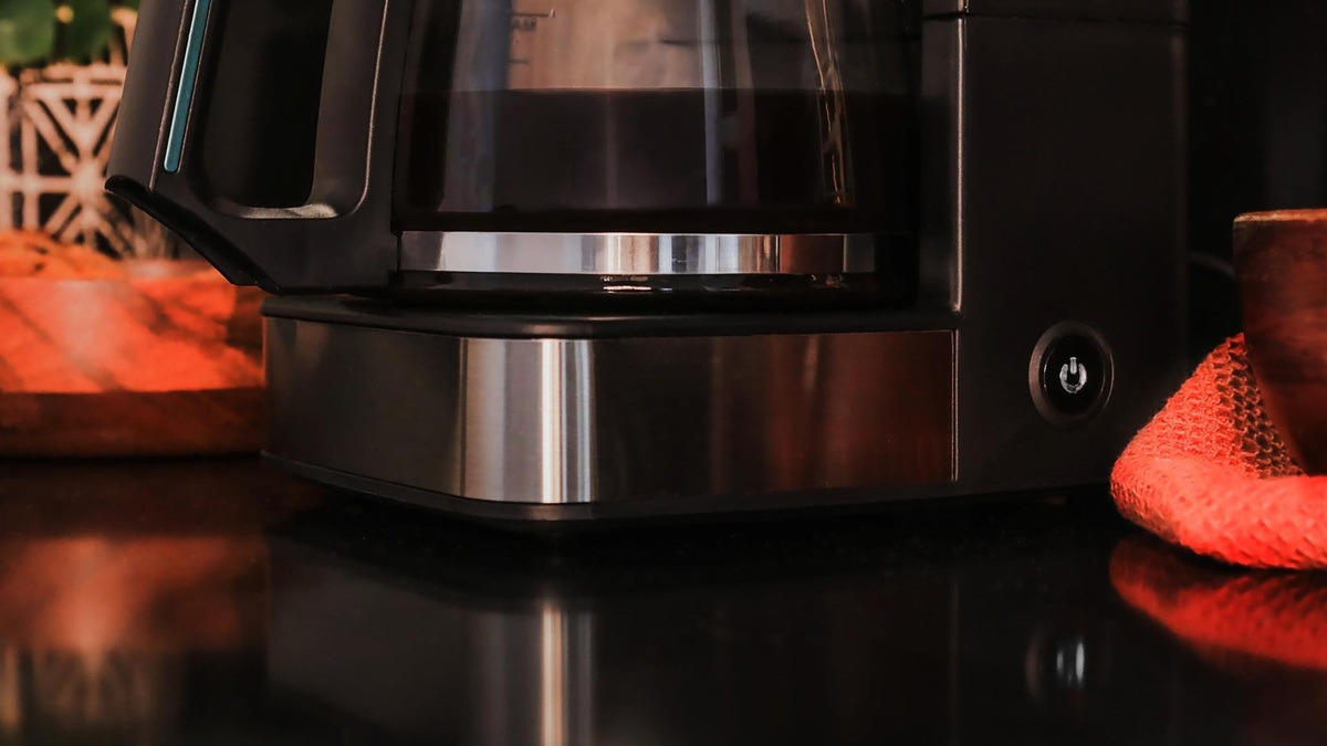 Zbliżenie ekspresu do kawy. Czarny i srebrny, ze szklanym dzbankiem pełnym kawy. Widoczny przycisk zasilania. Czerwona szmatka i drewniany przedmiot w pobliżu.