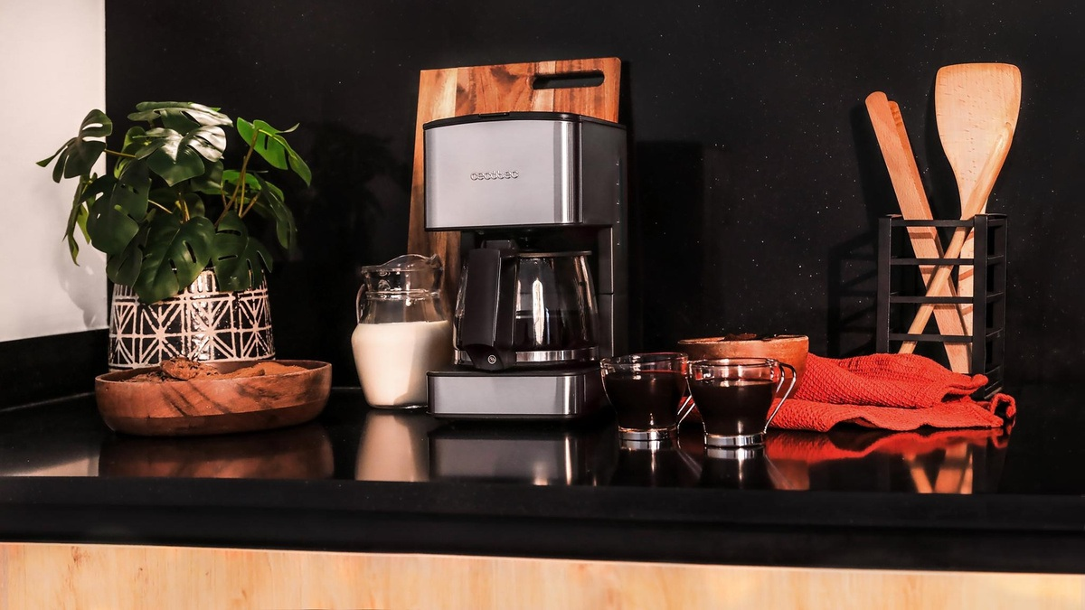 Ekspres do kawy, filiżanki kawy, drewniane przybory, roślina, dzbanek mleka i drewniana miska na czarnym blacie.