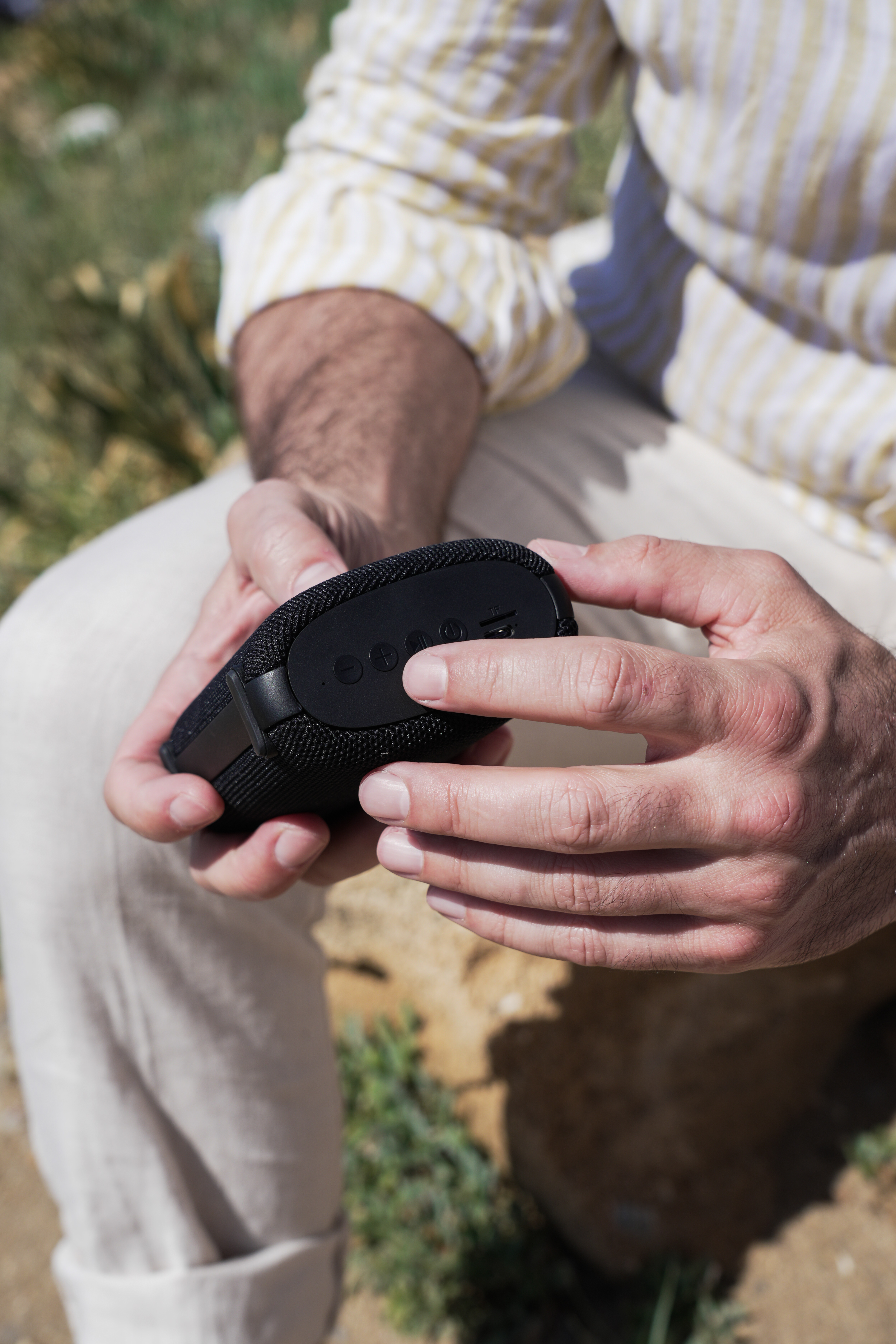 Osoba trzyma czarny głośnik z przyciskami. Osoba ma na sobie koszulę w paski i beżowe spodnie. Zielona trawa w tle.