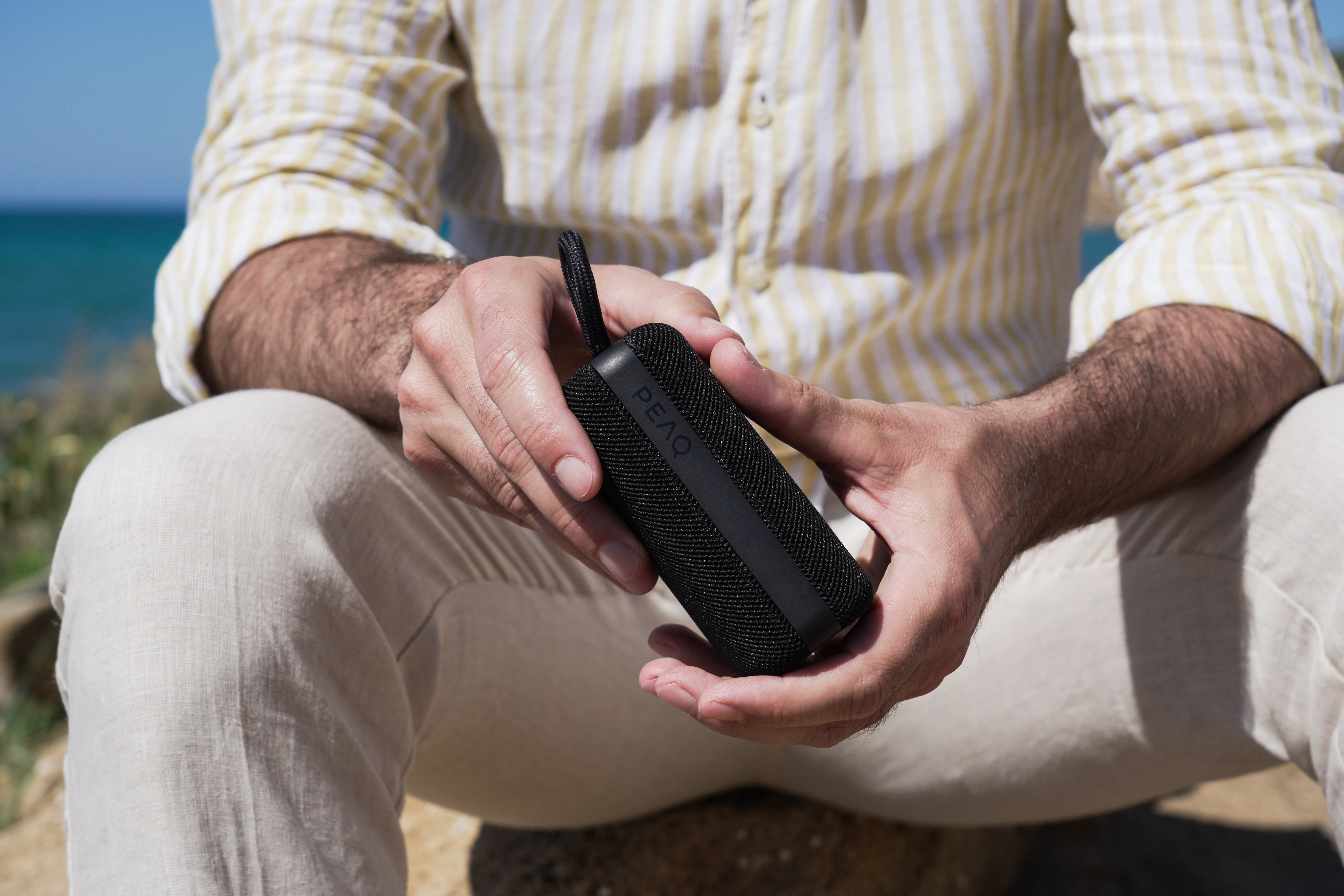 Osoba trzyma czarny głośnik PEAQ. Ma na sobie żółtą koszulę w paski i jasne spodnie. Tło to błękitne morze i niebo.