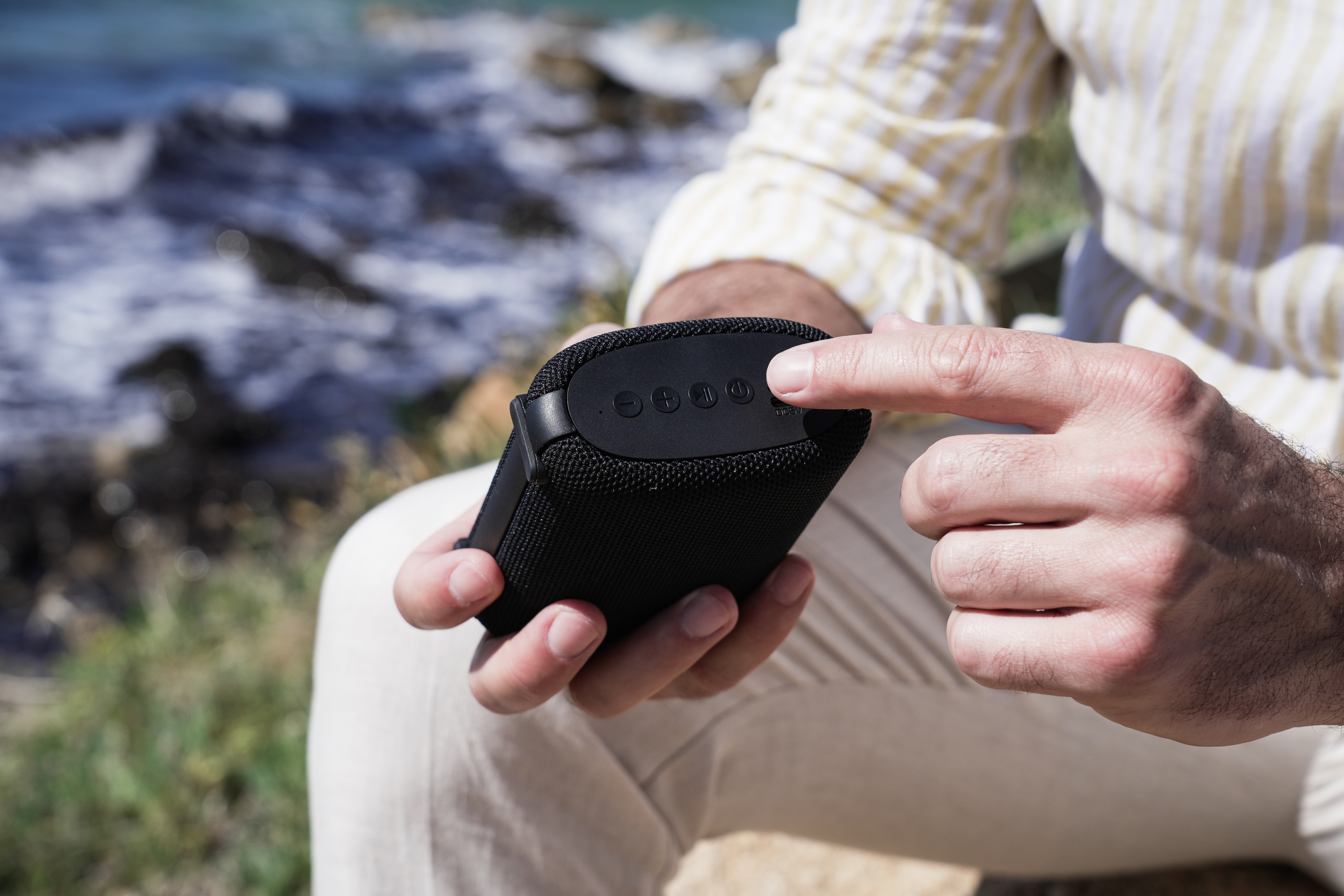 Dłoń naciska przycisk zasilania na czarnym głośniku. Tło to rozmyta woda i skały.