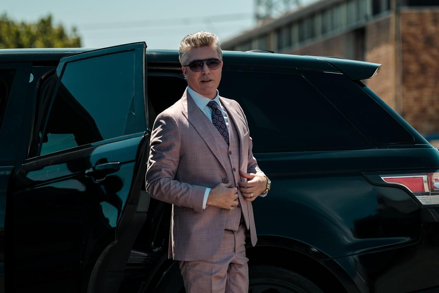Ein Mann in einem rosa Anzug und Sonnenbrille steht neben einem schwarzen SUV. Der Mann hält seine Jacke fest.