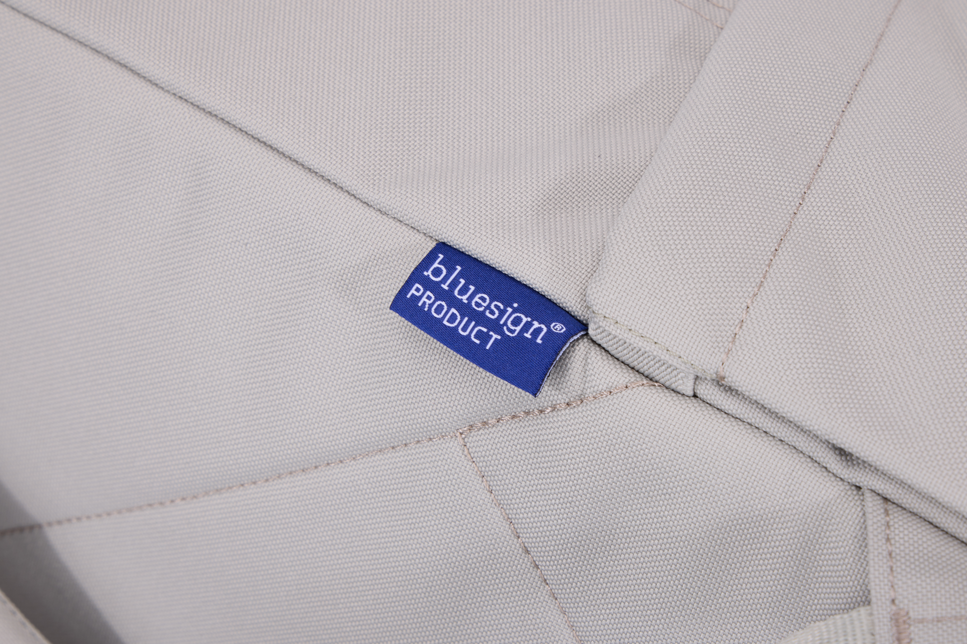 Mavi bir Bluesign ürün etiketi, açık gri bir kumaşın üzerinde. Kumaşın dikişleri ve dokulu bir yüzeyi var.