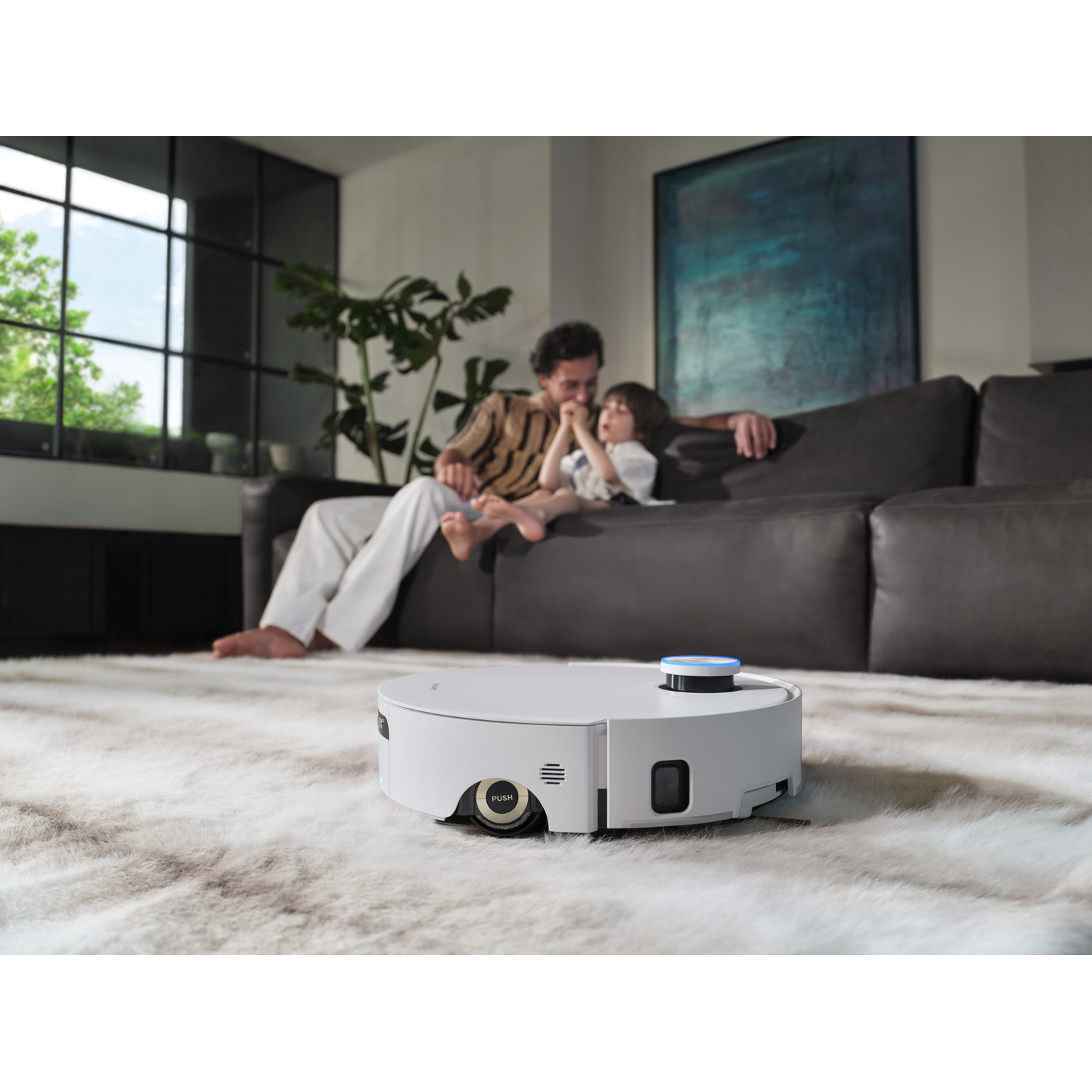 Een witte robotstofzuiger staat op een kleed. Een man en kind zitten op een bank op de achtergrond. Een raam is aan de linkerkant.