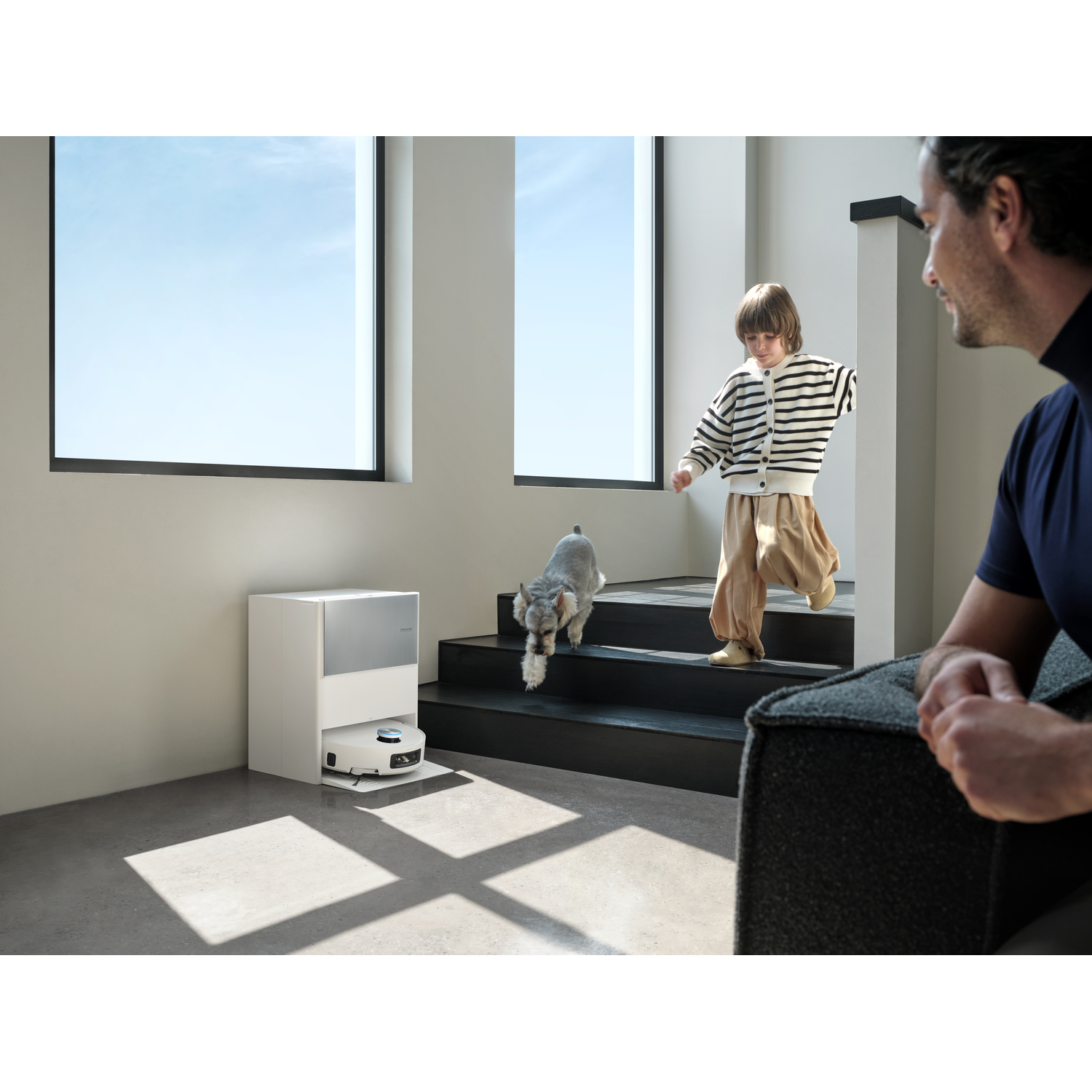 Een kind, een hond en een man zijn in een huis. Het kind rent de trap af. Een robotstofzuiger maakt schoon. Een man zit op een bank en glimlacht.