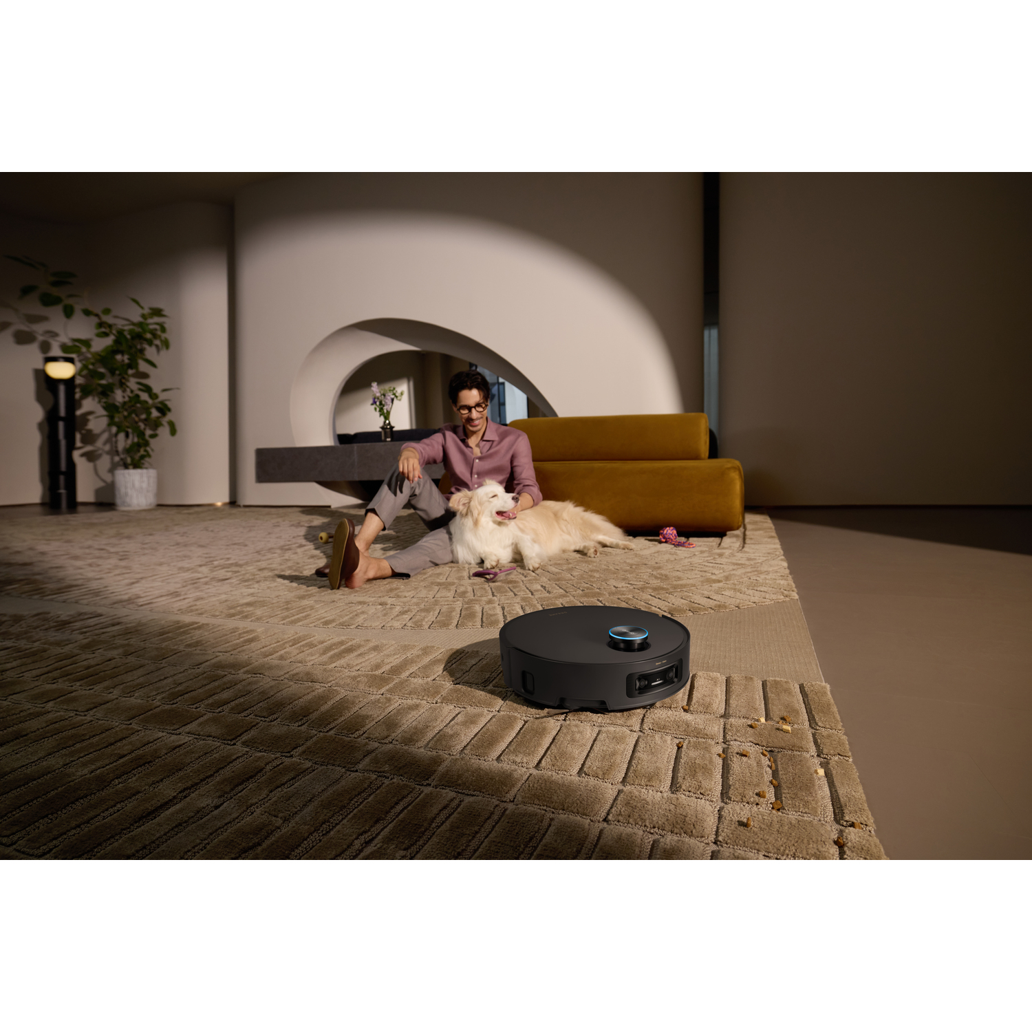 A man sits with a dog on a rug. A robotic vacuum cleaner is in the foreground. A yellow sofa and a plant pot are in the background.