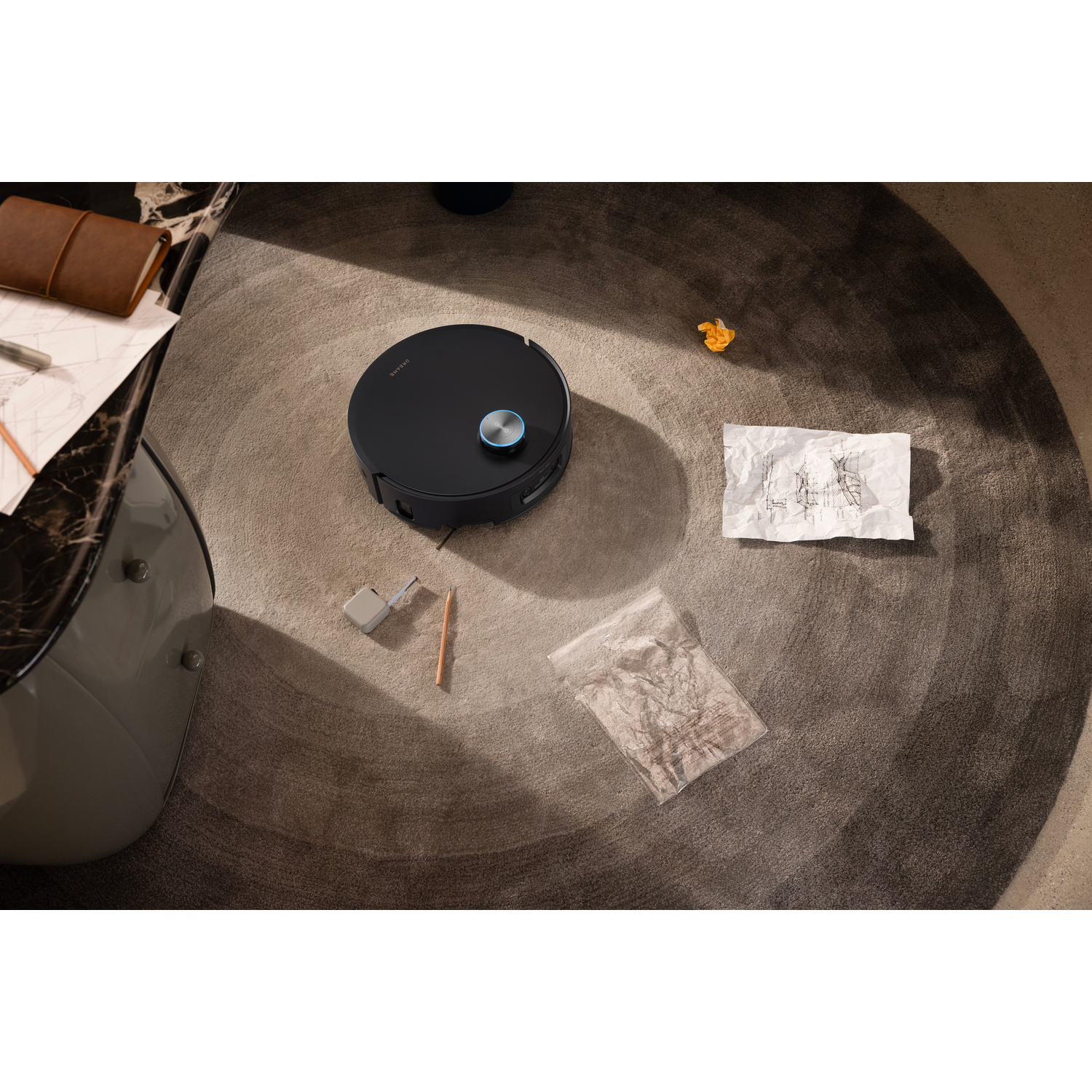 A black robot vacuum cleaner on a round carpet. Scattered around are a notepad, a pencil, crumpled paper, and a plastic bag.