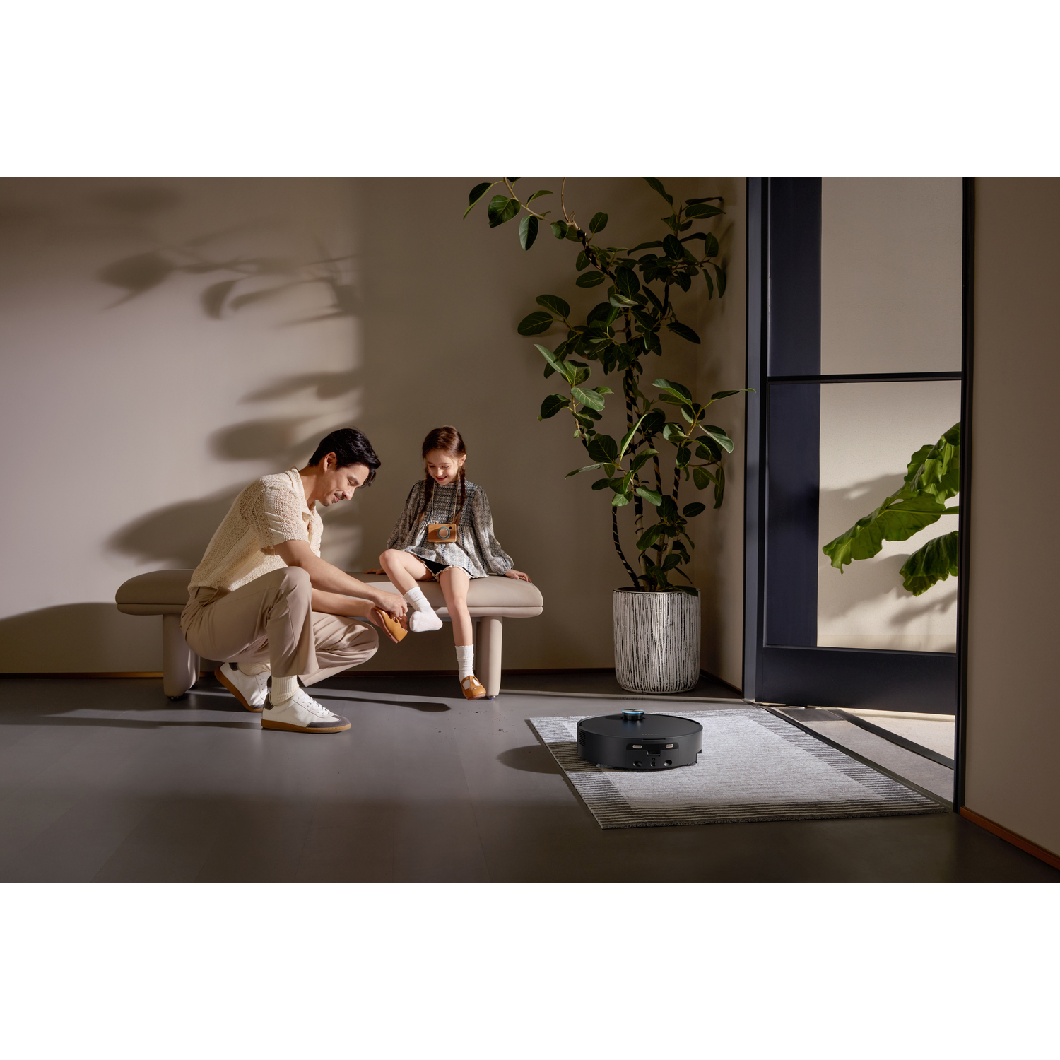 A man puts on a shoe for a girl on a bench. A robot vacuum cleaner sits on a rug. A large plant stands nearby.