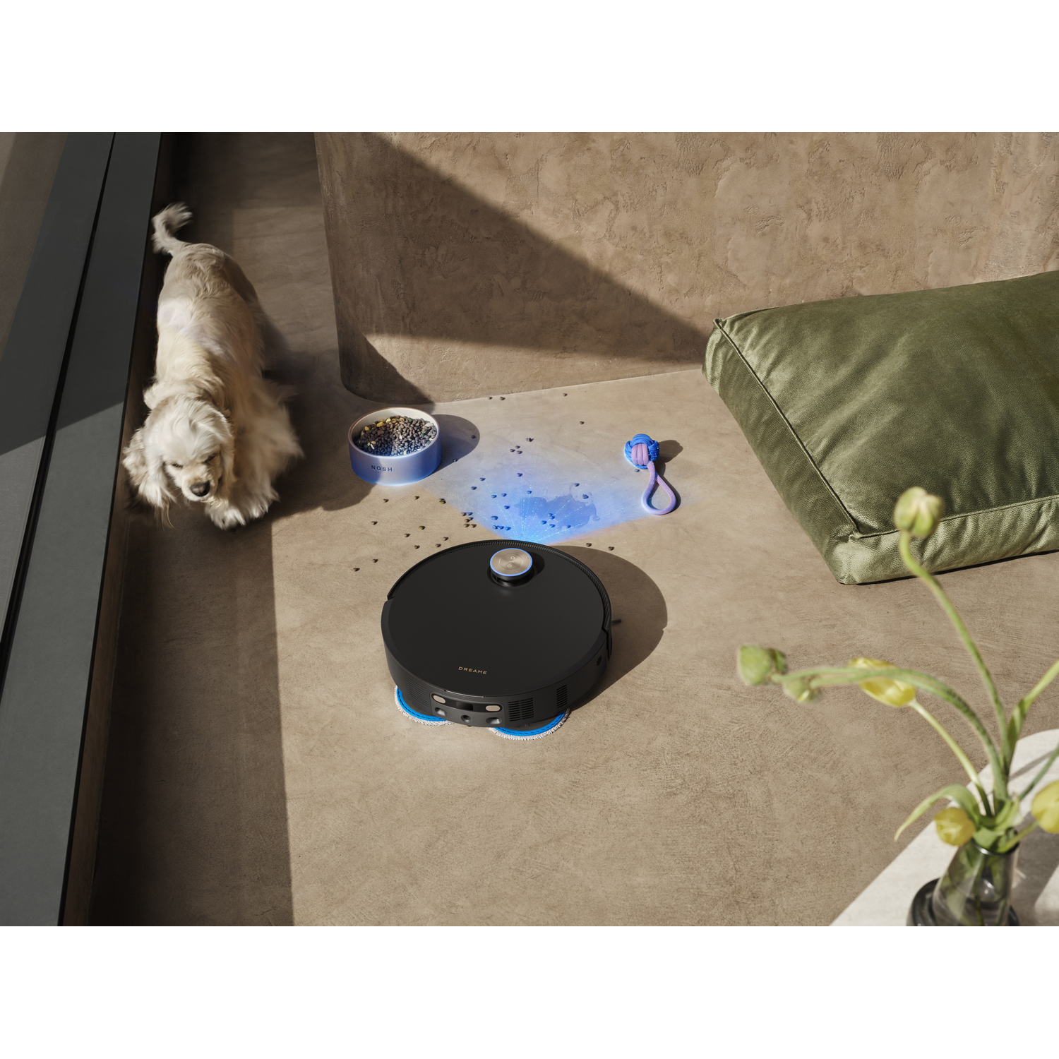 A black robot vacuum cleaner on a concrete floor. A dog, a pillow, and a vase with flowers are also in the scene.