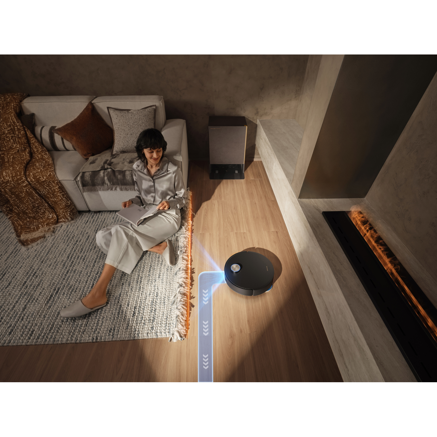 A woman sits with a book as a robot vacuum cleans the floor. A couch, rug, and fireplace are in the background.
