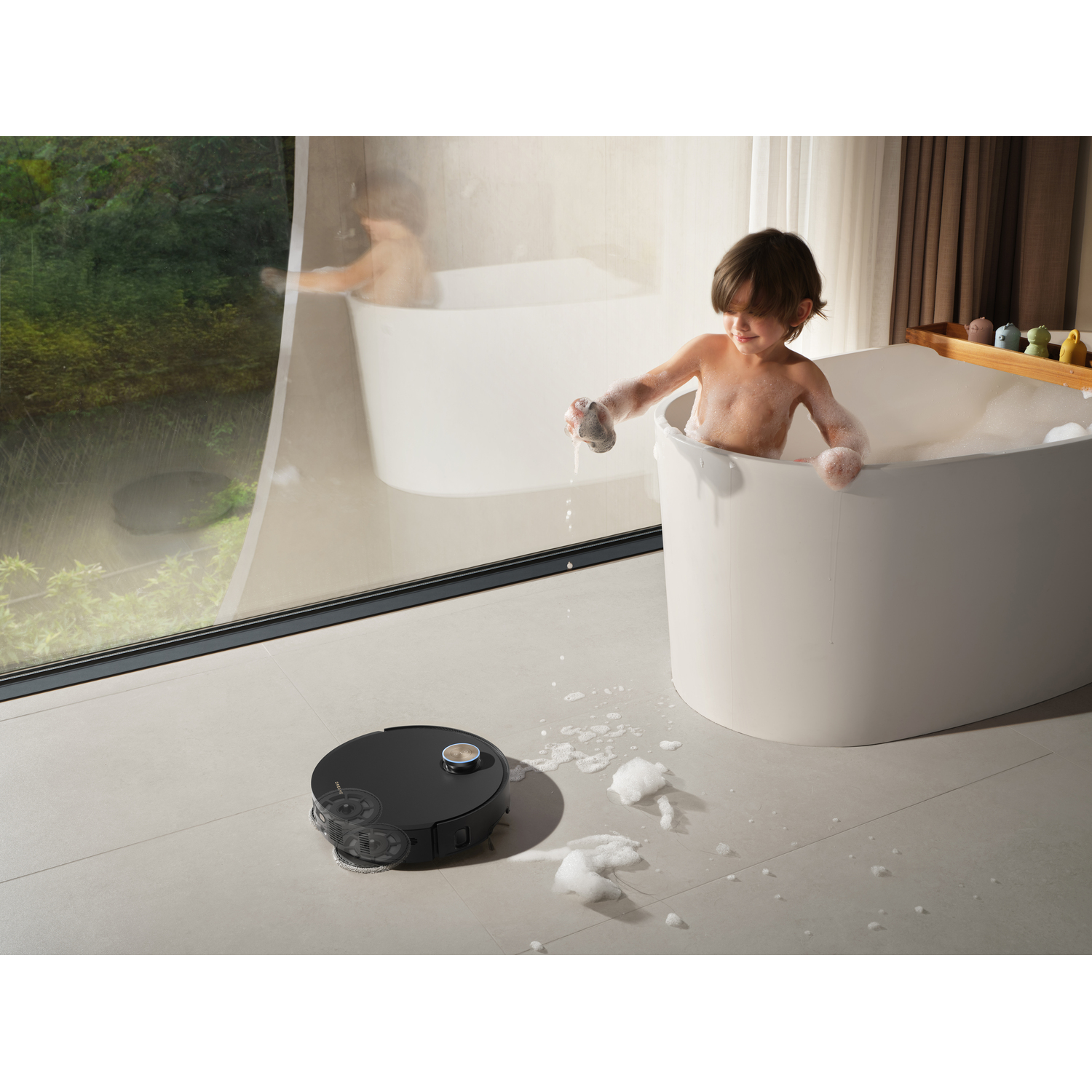 A child in a white bathtub plays with a sponge, surrounded by foam. A black robot vacuum cleans the floor nearby.