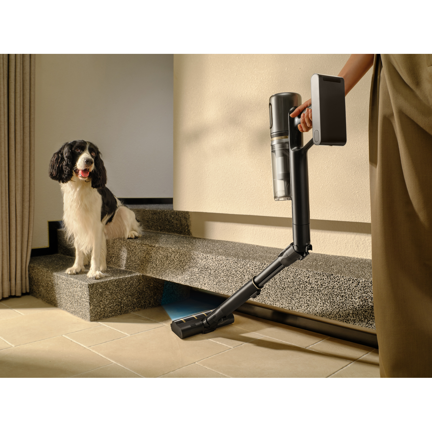 A black and white dog sits on steps as a person uses a vacuum cleaner. The vacuum is black and the floor is tiled.
