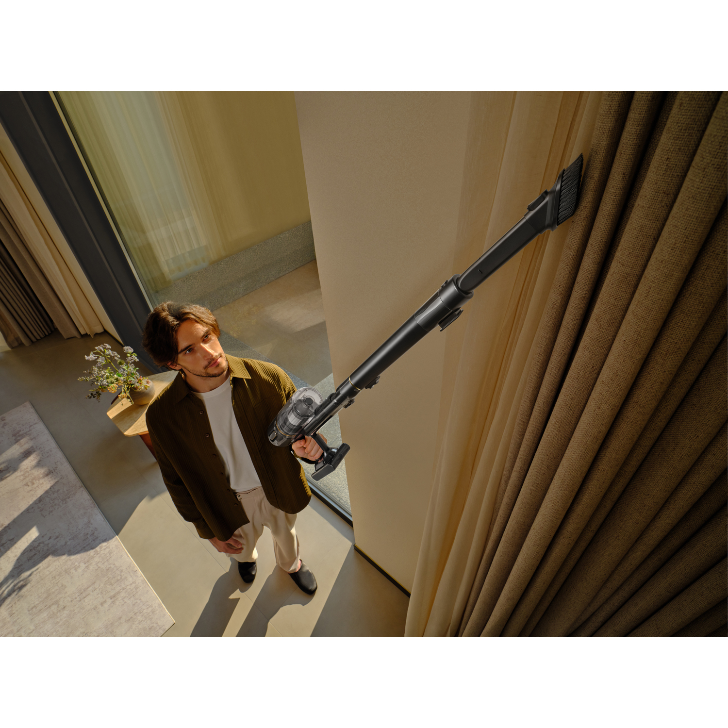 A man uses a vacuum cleaner on a beige curtain. He is wearing a green shirt and white pants. The room has a window.