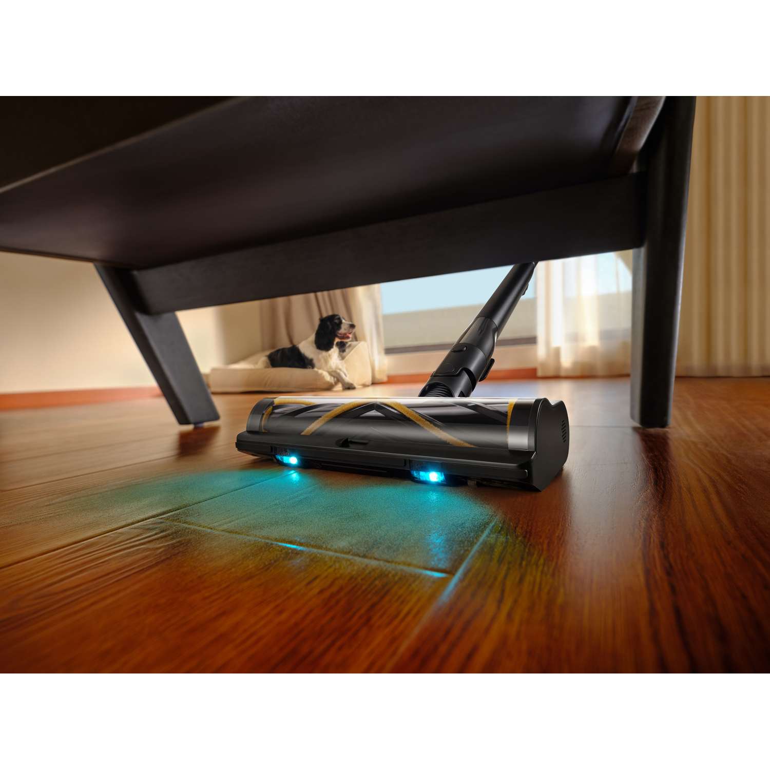 A black vacuum cleaner cleaning under a chair. A dog lies on a bed in the background. The floor is hardwood.