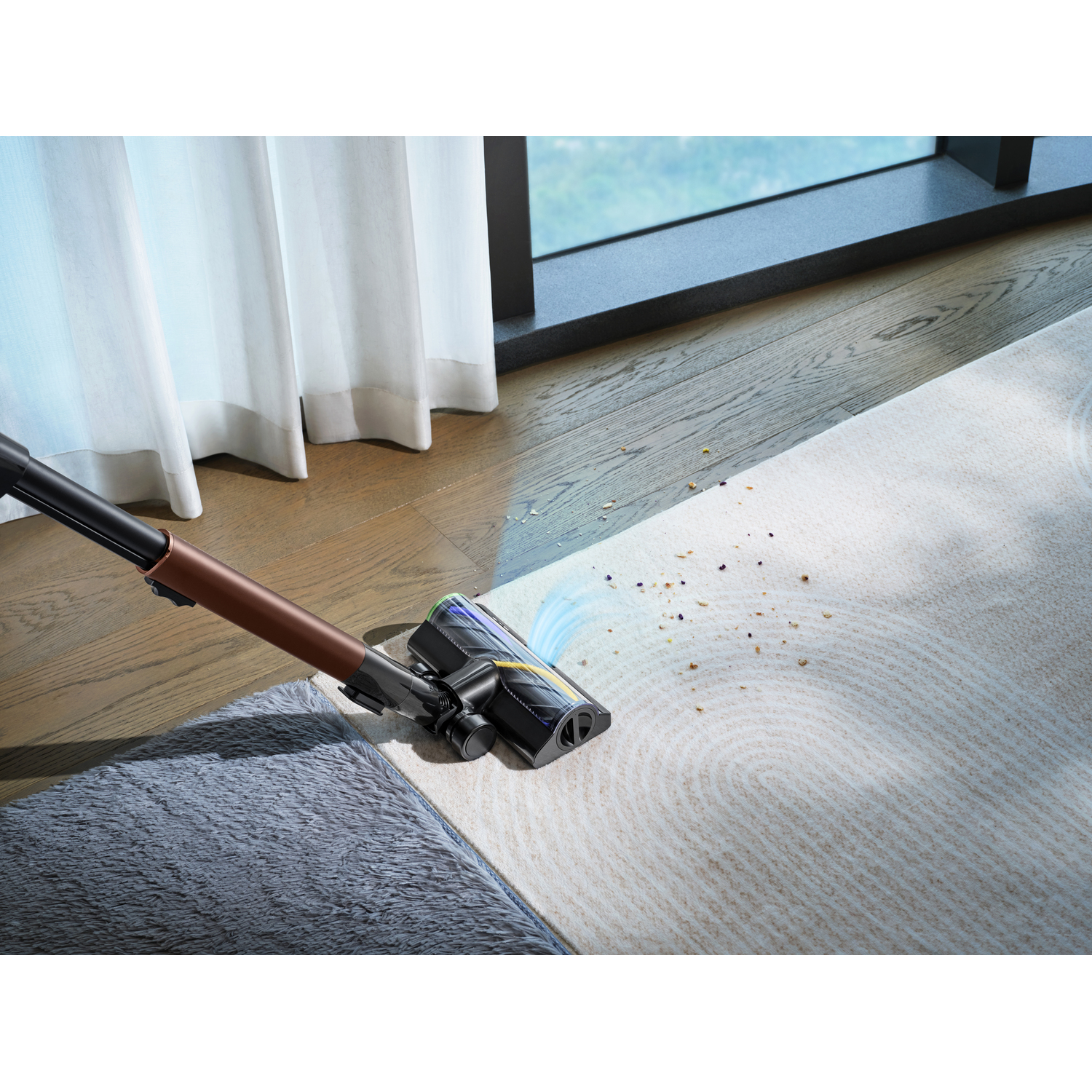 A cordless vacuum cleaner is cleaning a rug. Dust and debris are visible. A window and curtain are in the background.