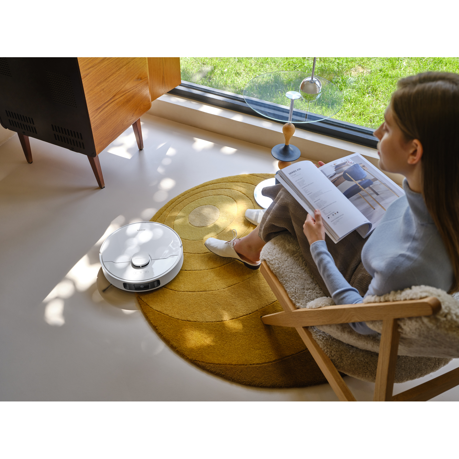 A woman reads while a robotic vacuum cleans. A yellow rug and a wooden cabinet are in the background. Sunlight streams through the window.