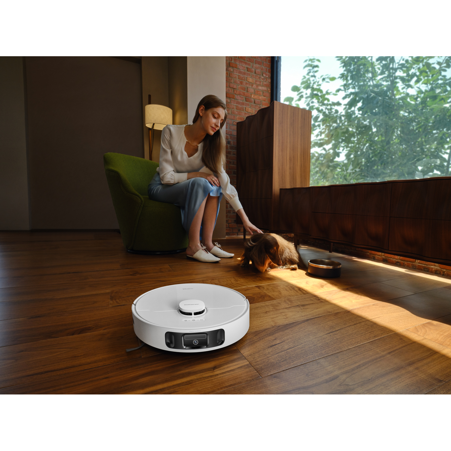 A woman sits near a window, petting a dog. A robotic vacuum cleaner is on the wooden floor. A lamp and cabinet are visible.