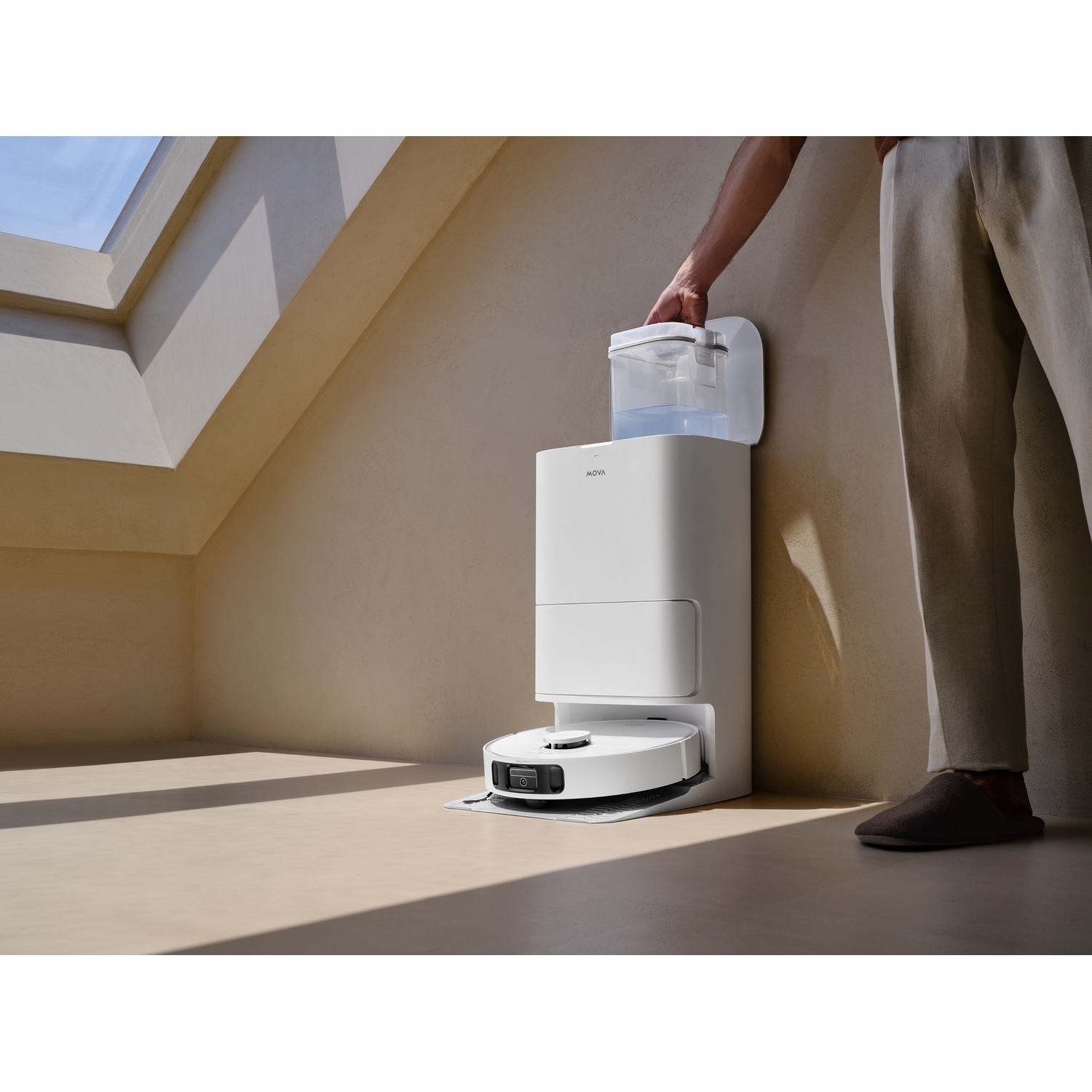 A white robot vacuum cleaner stands next to a white charging station. A person is placing a water tank on the station.