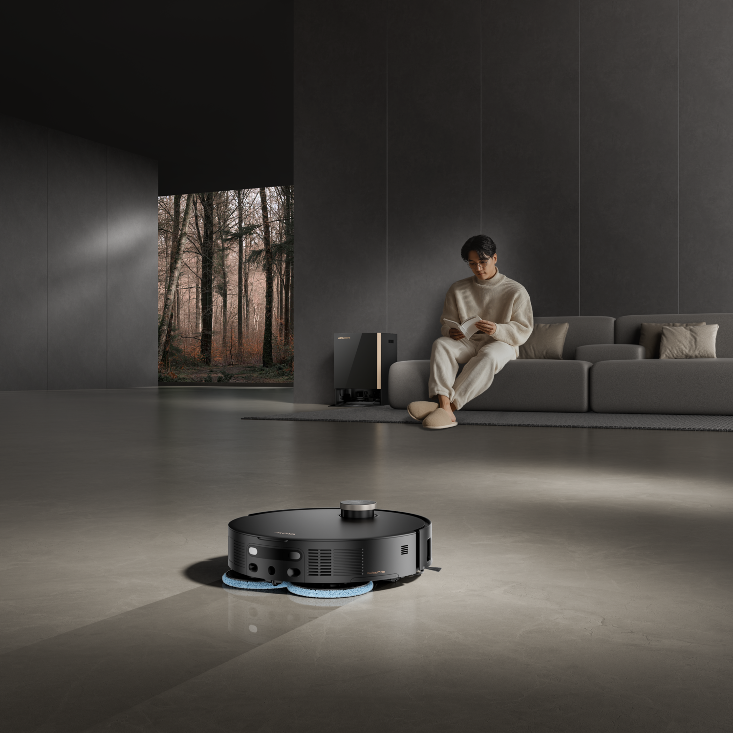 Un homme lit sur un canapé tandis qu'un aspirateur robot nettoie le sol. Une vue sur une forêt est visible à travers une fenêtre.