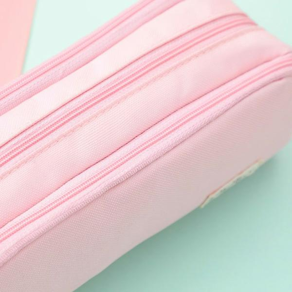 Zbliżenie na różowy piórnik z trzema zamkami błyskawicznymi. Tło jest jasnozielone i różowe.