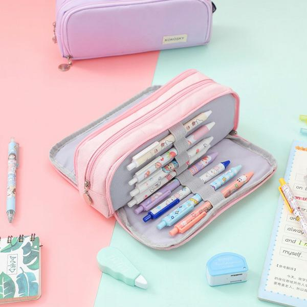 Różowy piórnik otwarty z długopisami i ołówkami w środku. Fioletowy piórnik na górze. Widoczne inne artykuły papiernicze.