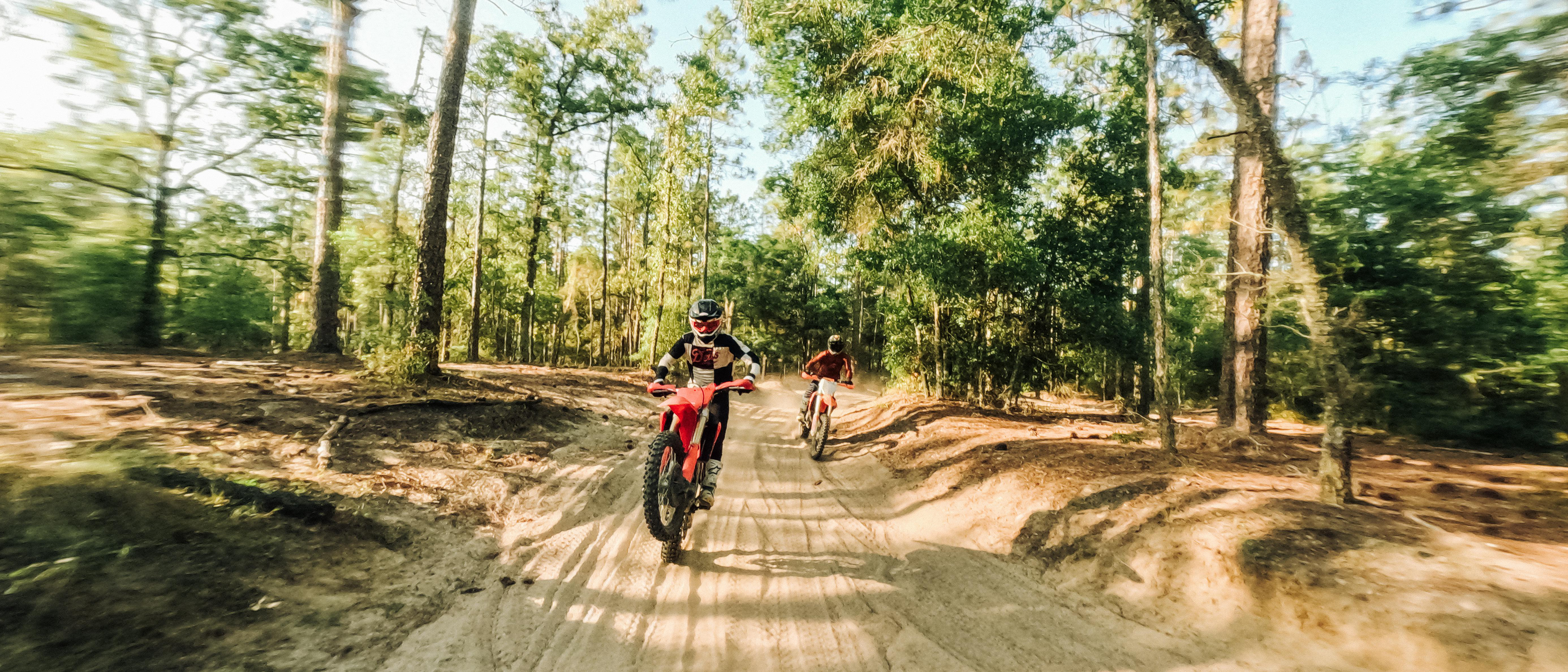 Zwei Motocross-Fahrer auf einem Feldweg im Wald. Ein Fahrer ist im Vordergrund, der andere weiter hinten. Bäume sind im Hintergrund.