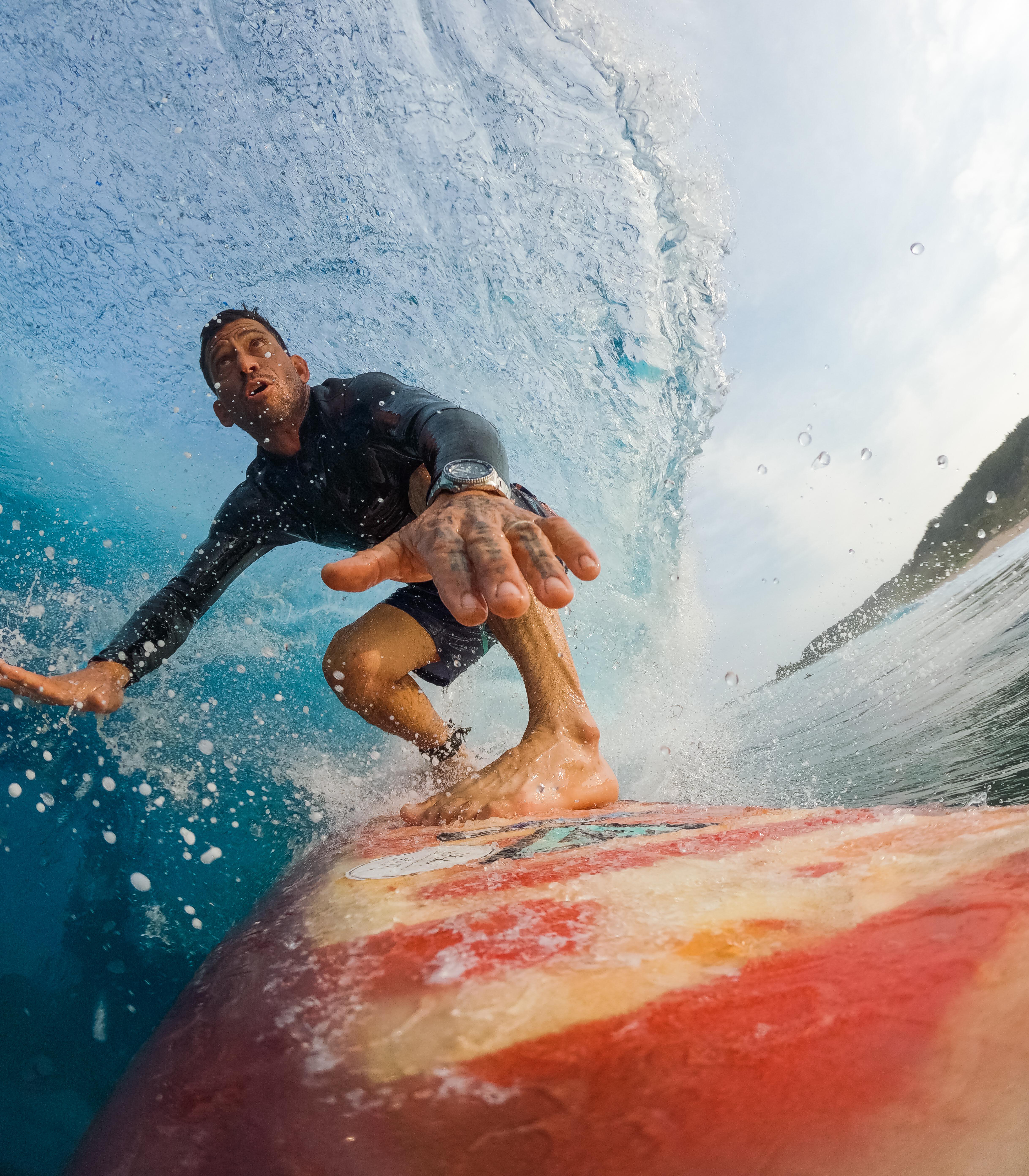 Ein Surfer reitet eine Welle. Er trägt einen schwarzen Neoprenanzug und ist auf einem rot-orangen Surfbrett.