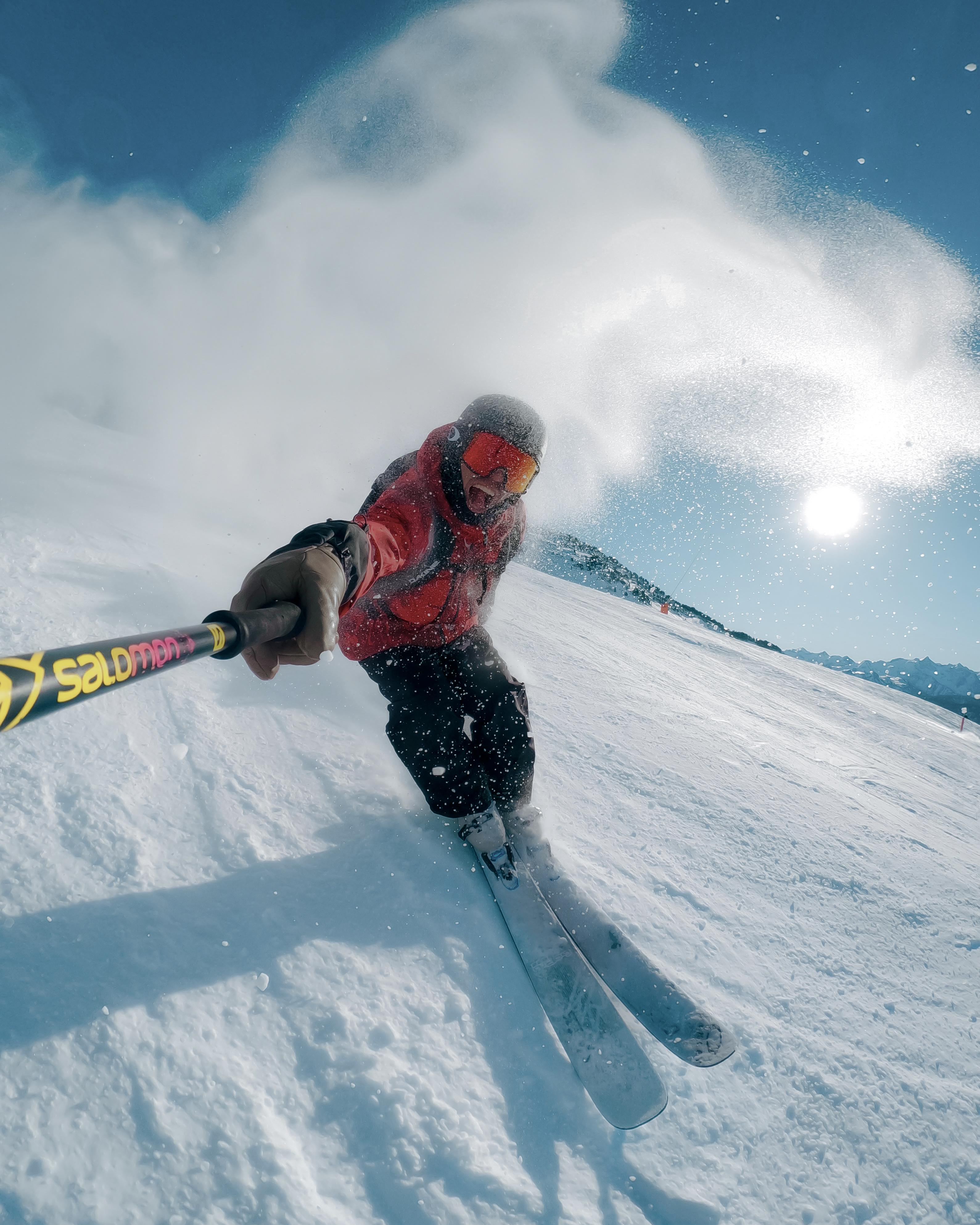 Skifahrer in roter Jacke fährt einen schneebedeckten Hang hinunter. Die Sonne scheint hell. Schnee spritzt in die Luft. Salomon-Stöcke sind sichtbar.