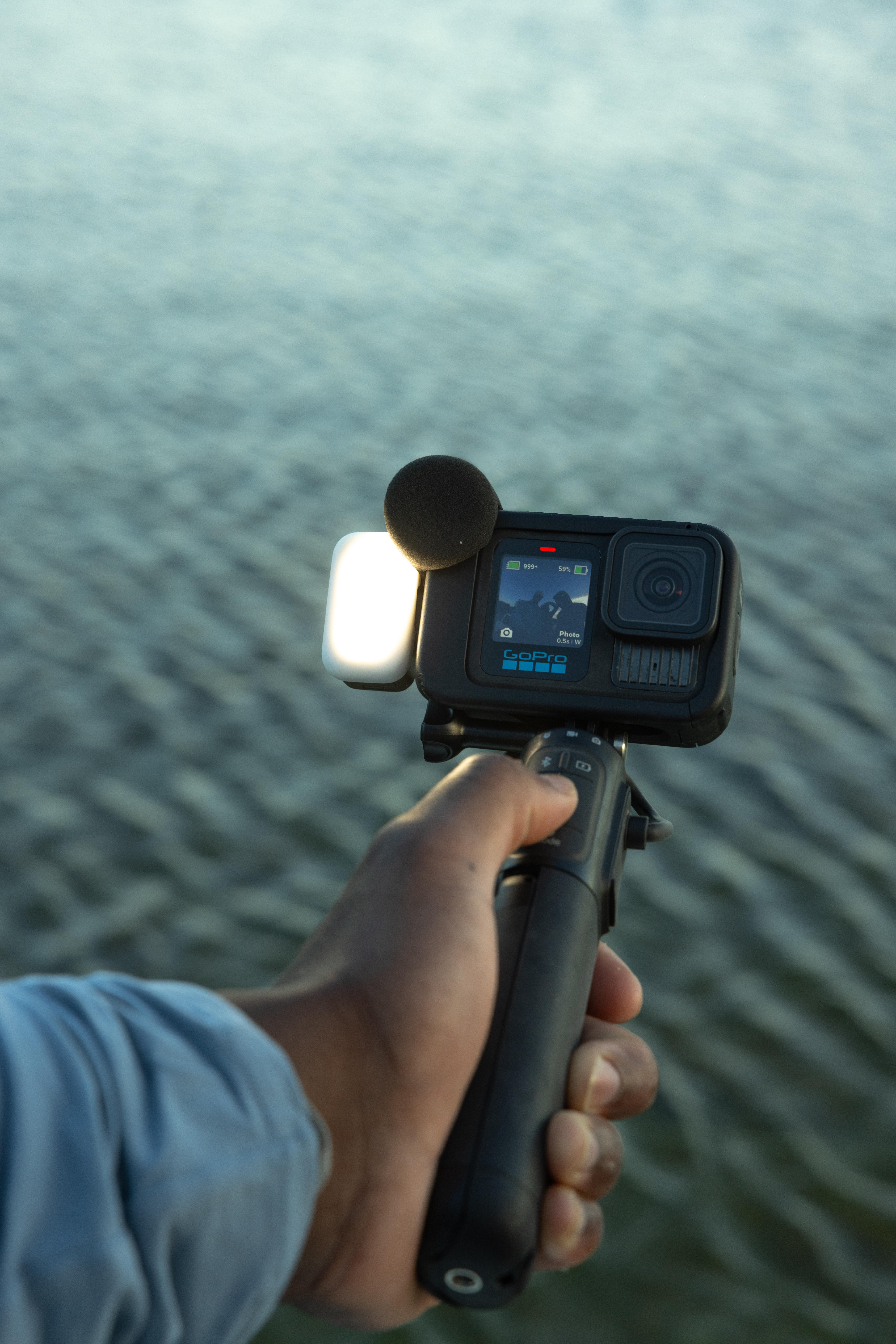 Eine Hand hält eine GoPro-Kamera an einem Selfie-Stick. Der Hintergrund ist ein Gewässer.