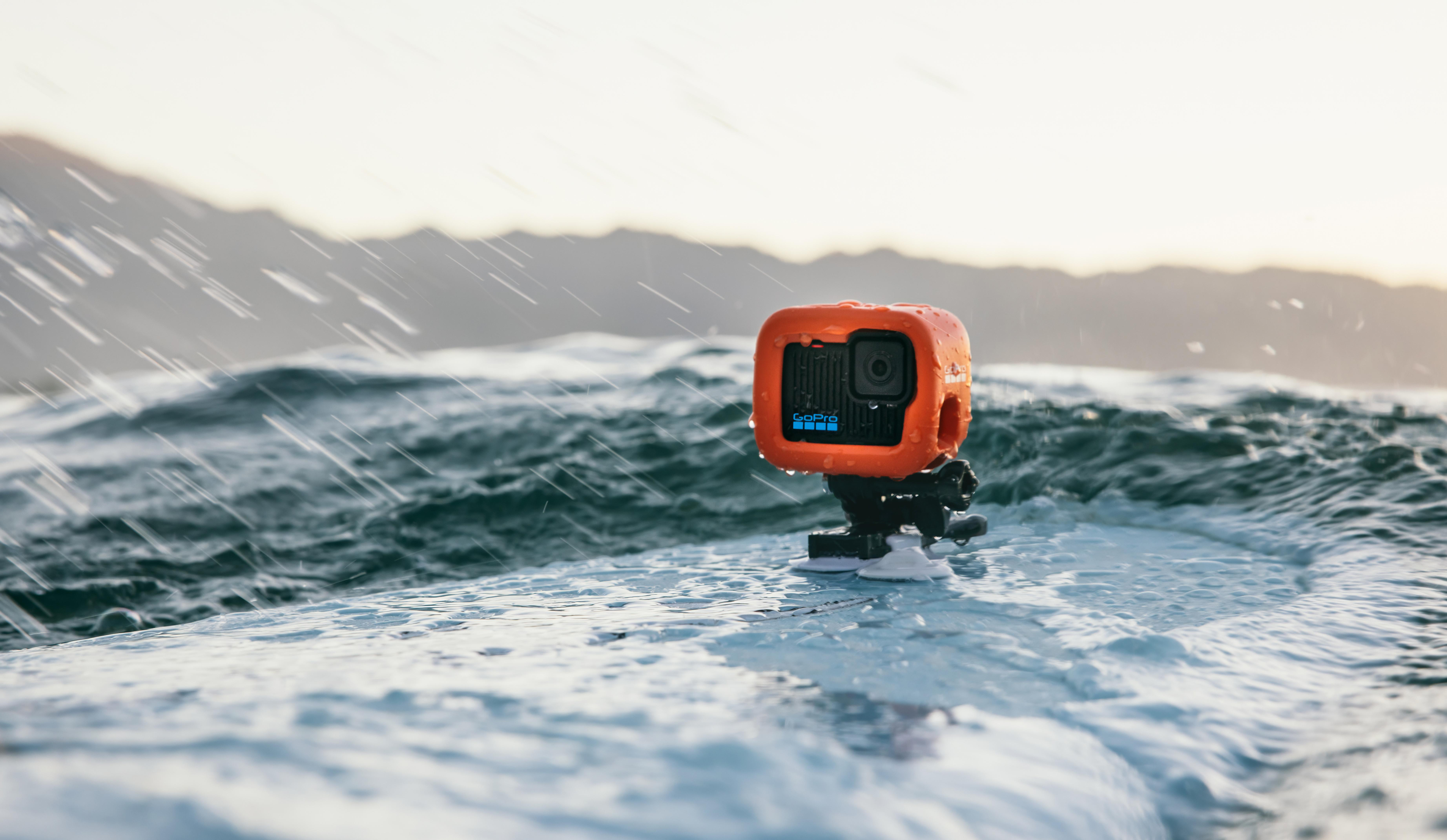 Eine orangefarbene GoPro-Kamera ist auf einem Surfbrett im Wasser montiert. Regen fällt. Berge im Hintergrund.