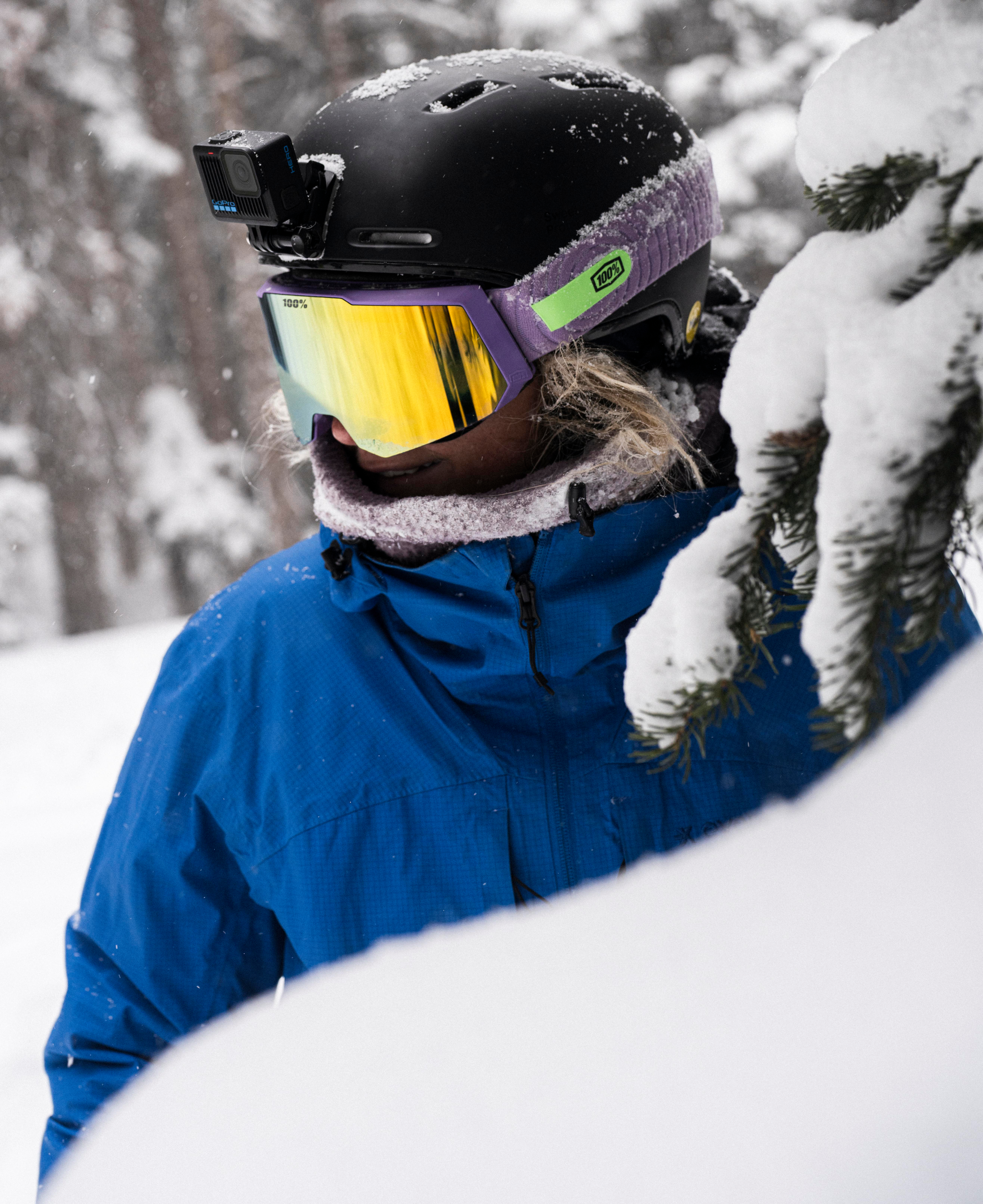 Person in blauer Jacke, Helm mit GoPro, Brille und Schal in einem verschneiten Wald. Vordergrund hat schneebedeckte Äste.