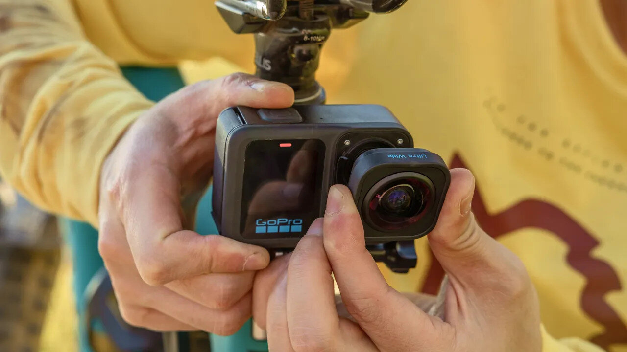 Osoba trzyma kamerę GoPro, regulując obiektyw. Kamera jest czarna i ma logo GoPro. Osoba ma na sobie żółtą koszulę.