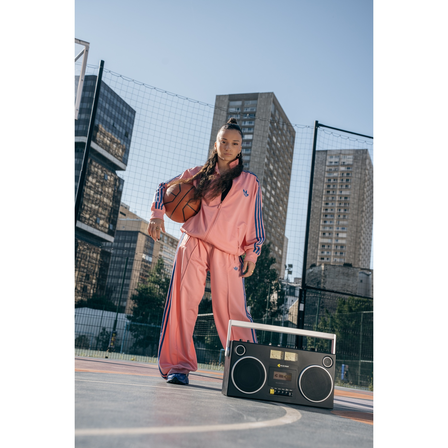 Una donna in tuta Adidas rosa tiene in mano un pallone da basket. È in piedi di fronte a una rete, con edifici sullo sfondo e un boombox.
