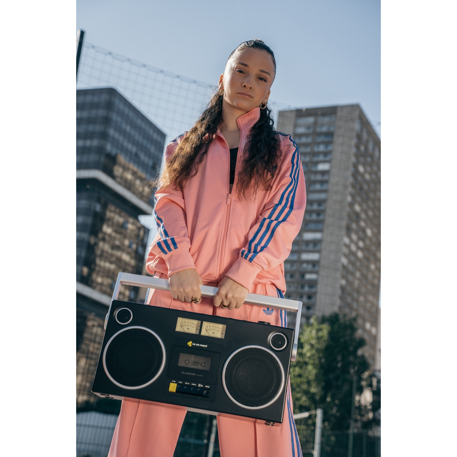 Una donna in tuta Adidas rosa tiene in mano un boombox. Edifici e rete sullo sfondo sotto un cielo blu. Guarda la fotocamera.