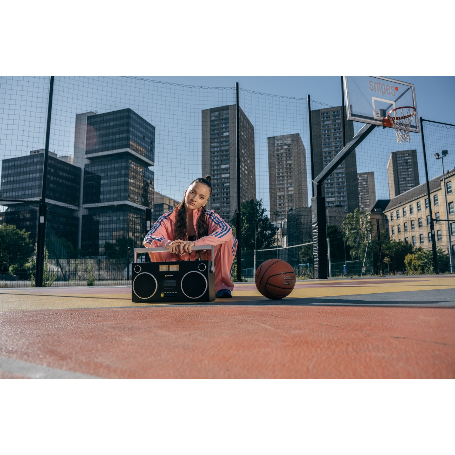 Una donna in tuta rosa è seduta su un campo da basket, appoggiata a un boombox. Un pallone e un canestro sullo sfondo, con edifici.