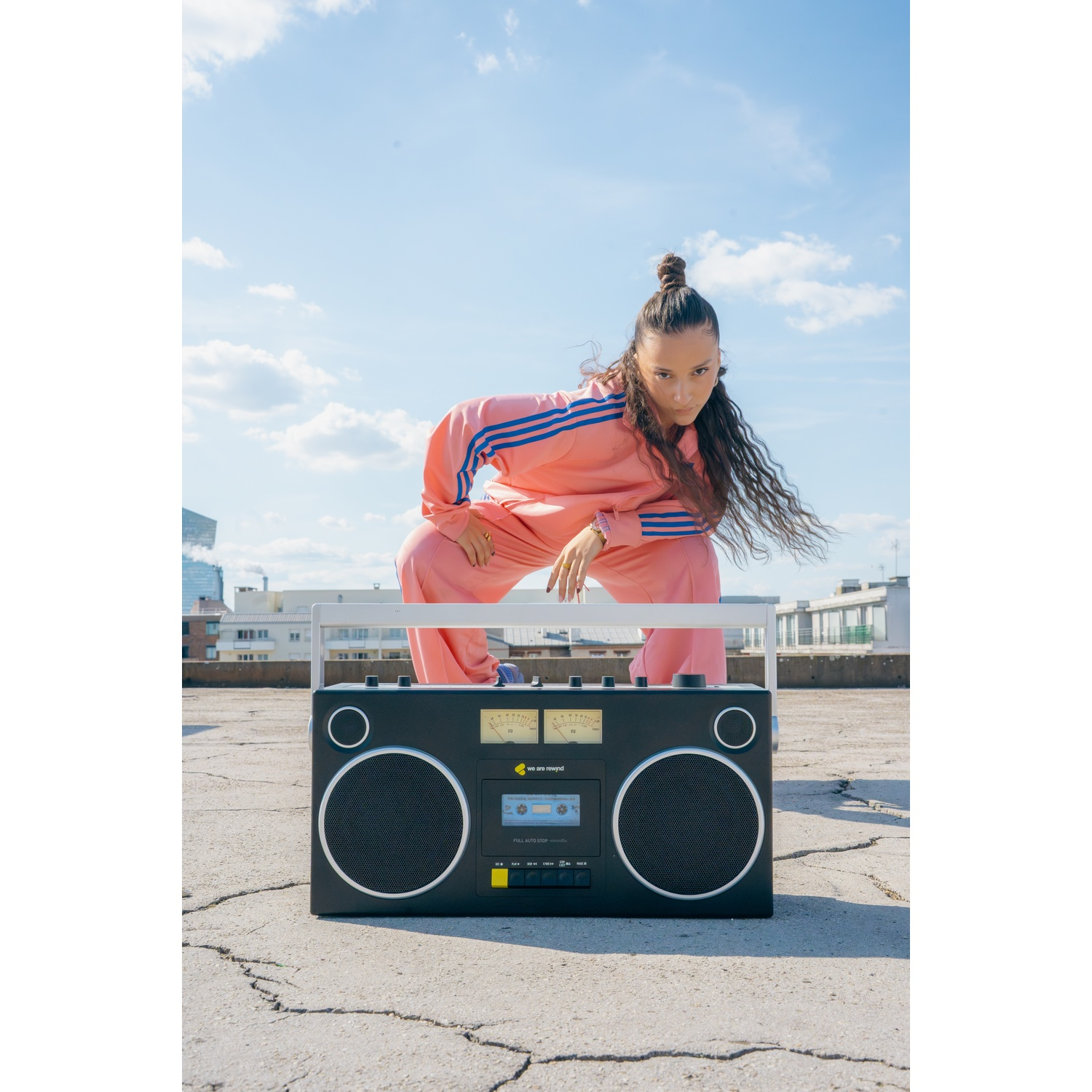Una donna in tuta rosa posa vicino a un grande boombox nero su un tetto. Cielo blu ed edifici sullo sfondo.