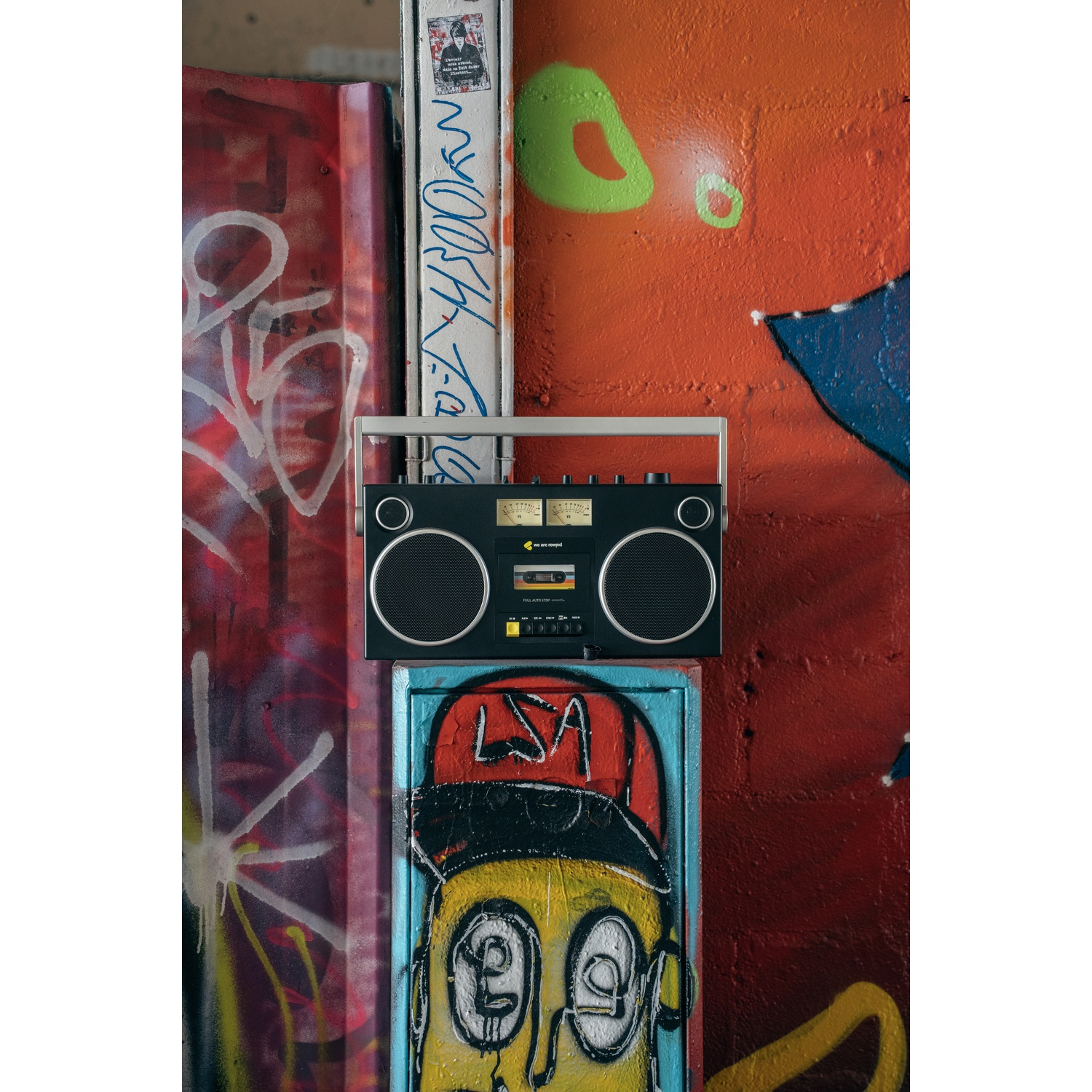 Un boombox è appeso a un muro con graffiti. Il muro è rosso e arancione con vari graffiti. Sotto il boombox c'è una faccia di cartone animato.