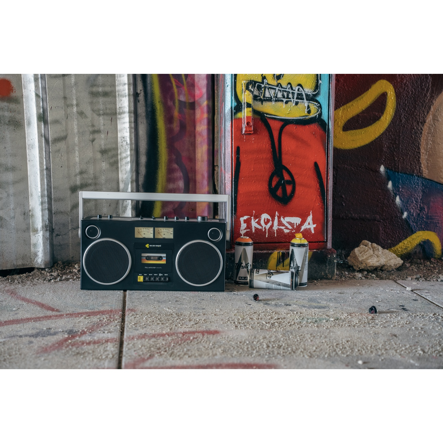 Un boombox nero è su cemento. Arte graffiti decora lo sfondo. Bombole spray accanto al boombox.