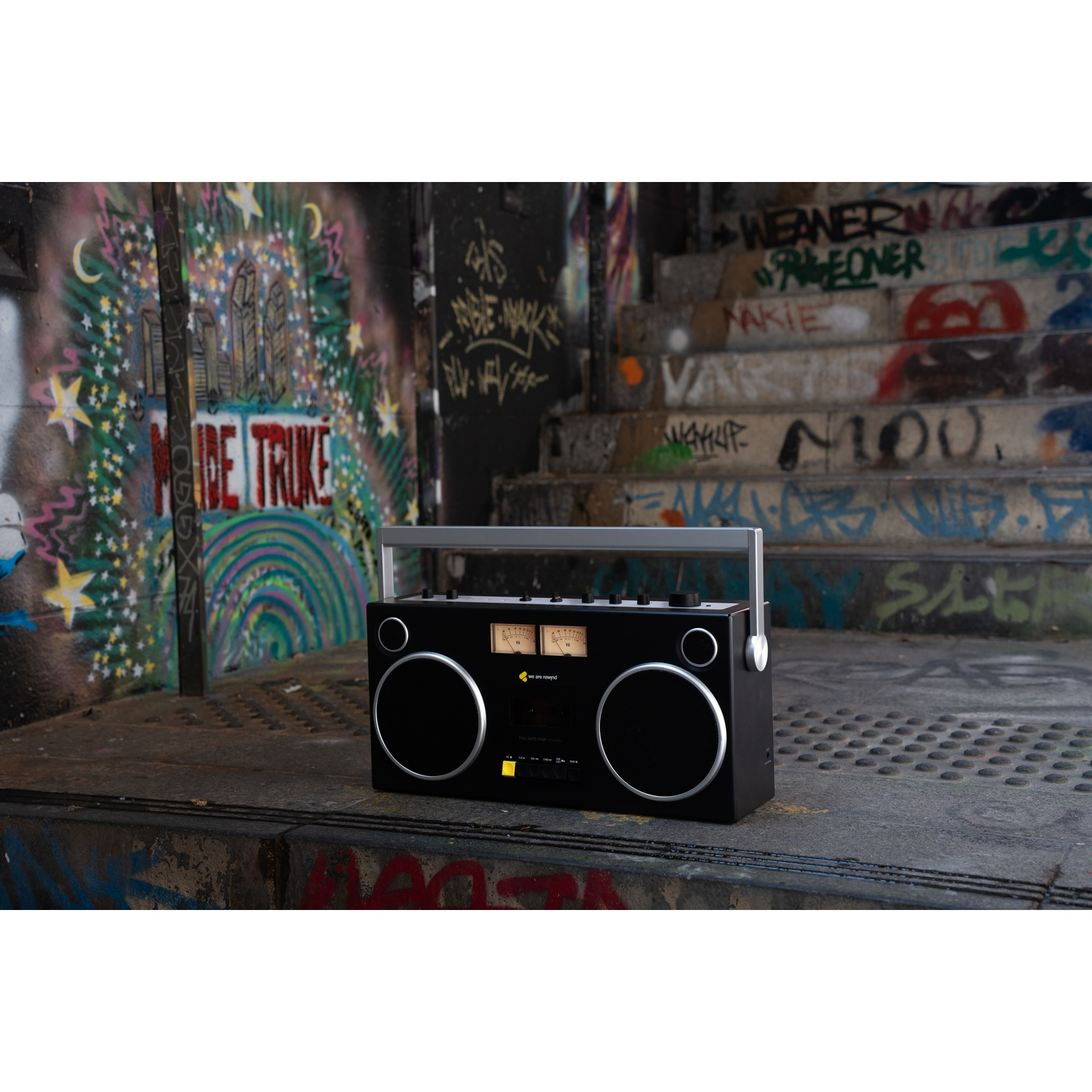 Un boombox nero con dettagli argentati è su una superficie metallica. I graffiti coprono il muro e le scale sullo sfondo.