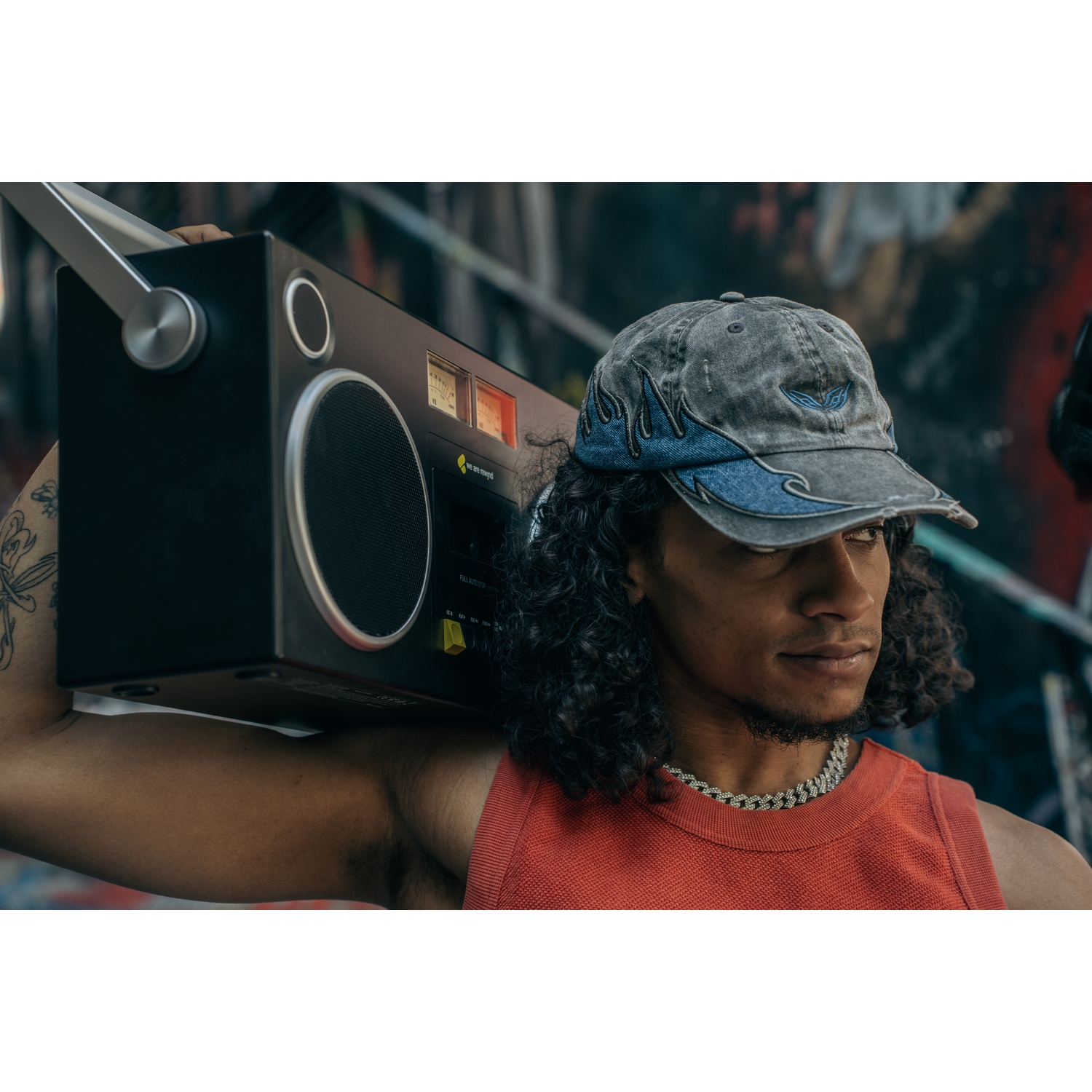 Uomo con capelli ricci porta un boombox nero sulla spalla, indossa un berretto e una maglia rossa, guarda altrove. Sfondo di graffiti.
