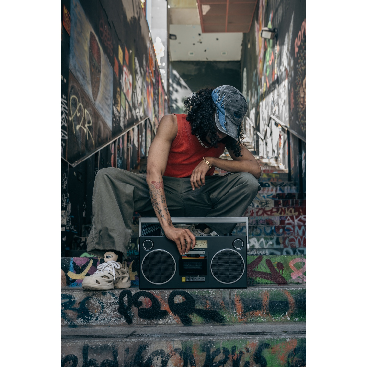 Una persona accovacciata con un boombox su gradini coperti di graffiti. Indossa canotta rossa, berretto e pantaloni verde oliva, mani vicino al boombox.