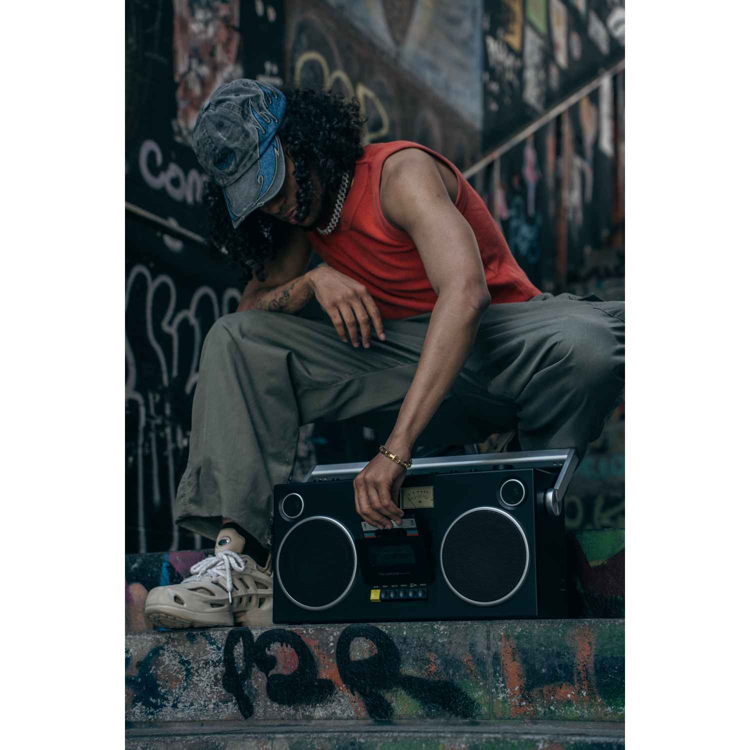 Un uomo con una maglietta rossa e un berretto è accovacciato vicino a un boombox, davanti a un muro di graffiti.
