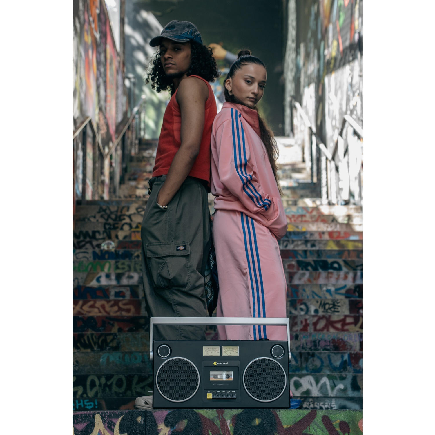 Due persone schiena contro schiena su scale coperte di graffiti, un boombox davanti. Uno indossa rosso, l'altro rosa, entrambi rivolti verso la fotocamera.